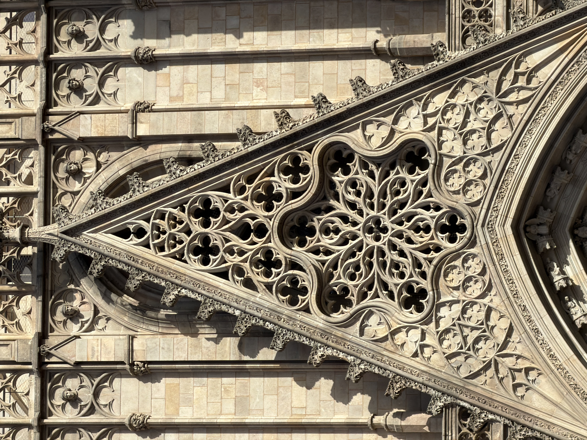 The image shows a detailed view of the facade of the Barcelona Cathedral, located in the Gothic Quarter of Barcelona, Spain. The central focus is a large, triangular Gothic window filled with intricate stone tracery. The tracery features a rose window design with multiple lobes and decorative elements resembling quatrefoils and trefoils. The stone itself is a light beige color, weathered and showing the effects of time.
Surrounding the window, the facade is constructed of stone blocks, with decorative architectural elements. To the left and right of the main triangular pediment, there are vertical sections of the facade with repeating arched niches. Within some of these niches, carved busts of human heads are visible, facing forward or slightly to the side. These appear to be aged stone sculptures.
The architectural style is clearly Gothic, characterized by pointed arches, ornate carvings, and the complex geometrical patterns in the window tracery. The sunlight casts strong shadows across the facade, highlighting the three-dimensional relief of the carvings and the depth of the window. The overall impression is one of grand historical architecture with rich detailing.