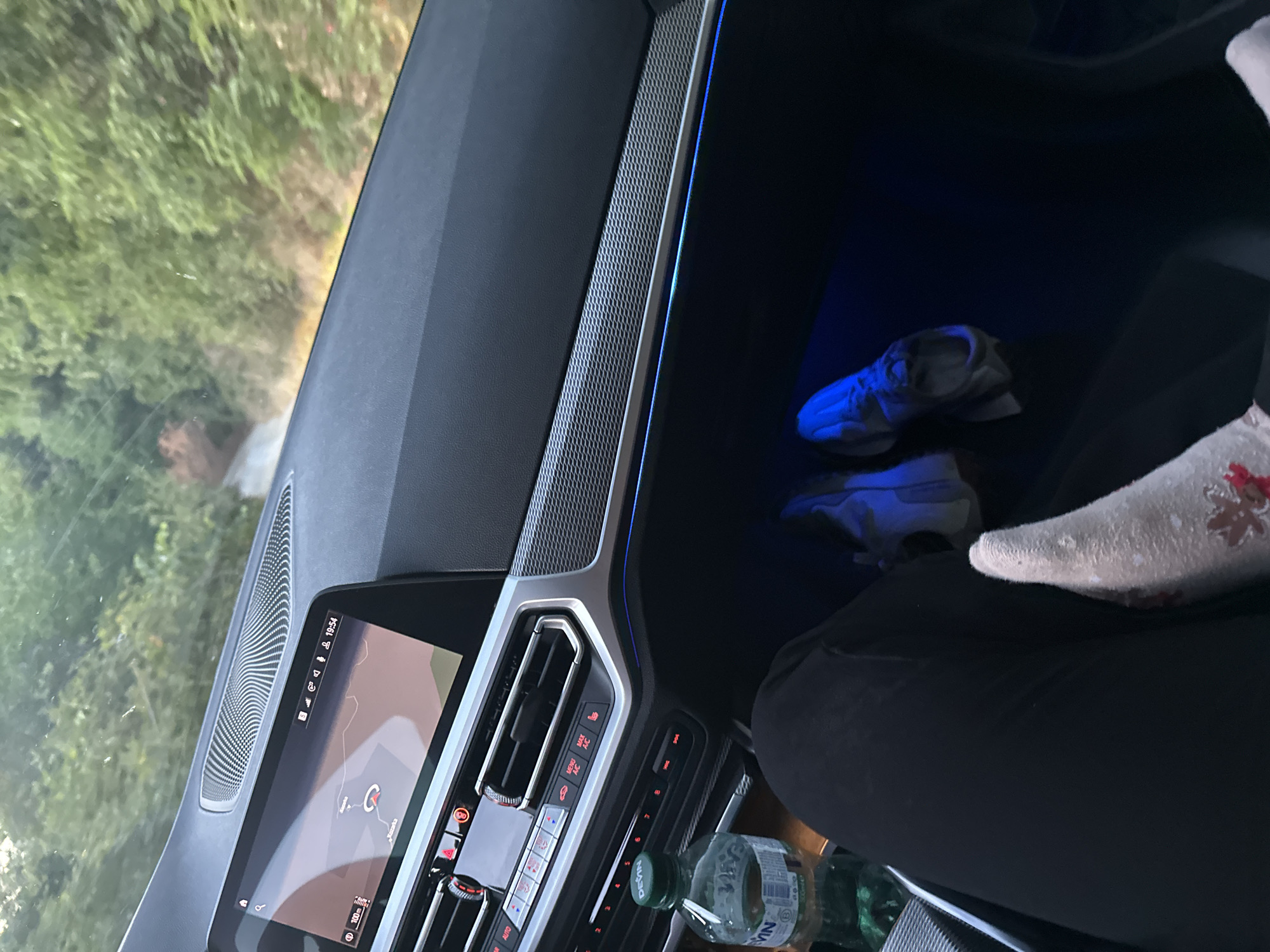 The image shows the interior of a car driving on a road near Kozarka, Bulgaria. The driver's perspective shows a dashboard with a large infotainment screen displaying a navigation map. The screen shows a road leading towards a point labeled "Kozarka." Outside the car, lush green trees line the road, indicating a rural or semi-rural setting. A bottle of water is visible on the center console, and a pair of sneakers rests on the floor, illuminated by ambient blue lighting. The overall scene suggests a journey through a scenic Bulgarian landscape.