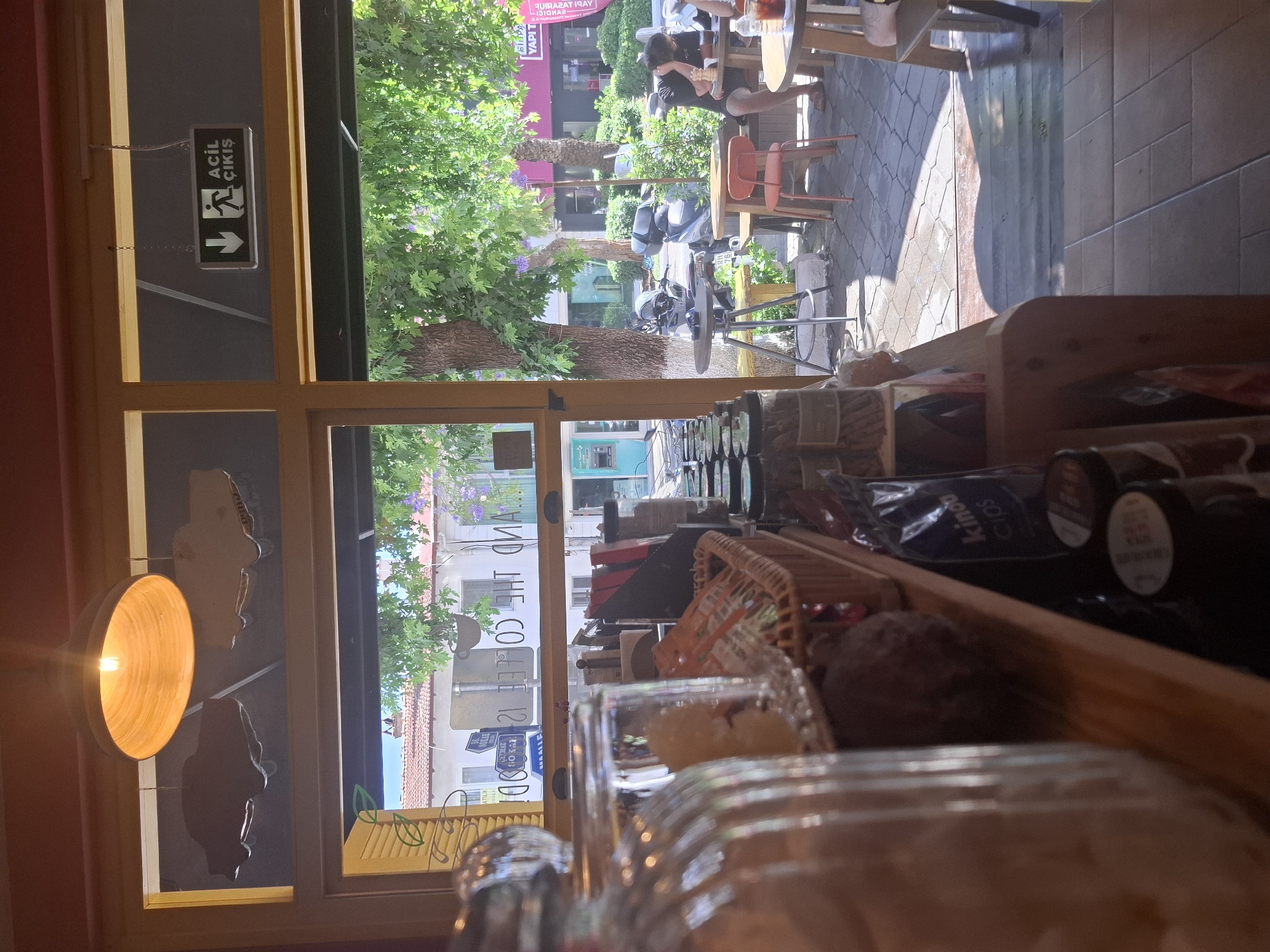 The image depicts the interior of a coffee shop in Fethiye, Turkey, looking out onto a sun-drenched outdoor seating area and street. In the foreground, a shelf displays various items, including glass jars with food items, a woven basket filled with packaged goods, and bags of coffee or snacks. A prominent glass jar filled with what appears to be dried goods is in the left foreground, blurred.
Through the large windows, the outdoor seating area is visible. Several round tables with chairs are arranged on a paved patio. One person is seated at a table on the right, facing away from the camera, appearing to be dining or resting. Trees with lush green foliage and some purple flowers provide shade. In the background, buildings with red-tiled roofs and shops line the street. A blue ATM machine is visible to the left.
Above the window, an illuminated exit sign with the text "ACIL ÇIKIŞ" (Emergency Exit in Turkish) is mounted, indicating the location is a public establishment. A decorative light fixture hangs from the ceiling, casting a warm glow. The overall impression is a bright, daytime scene in a relaxed, possibly Mediterranean, setting.