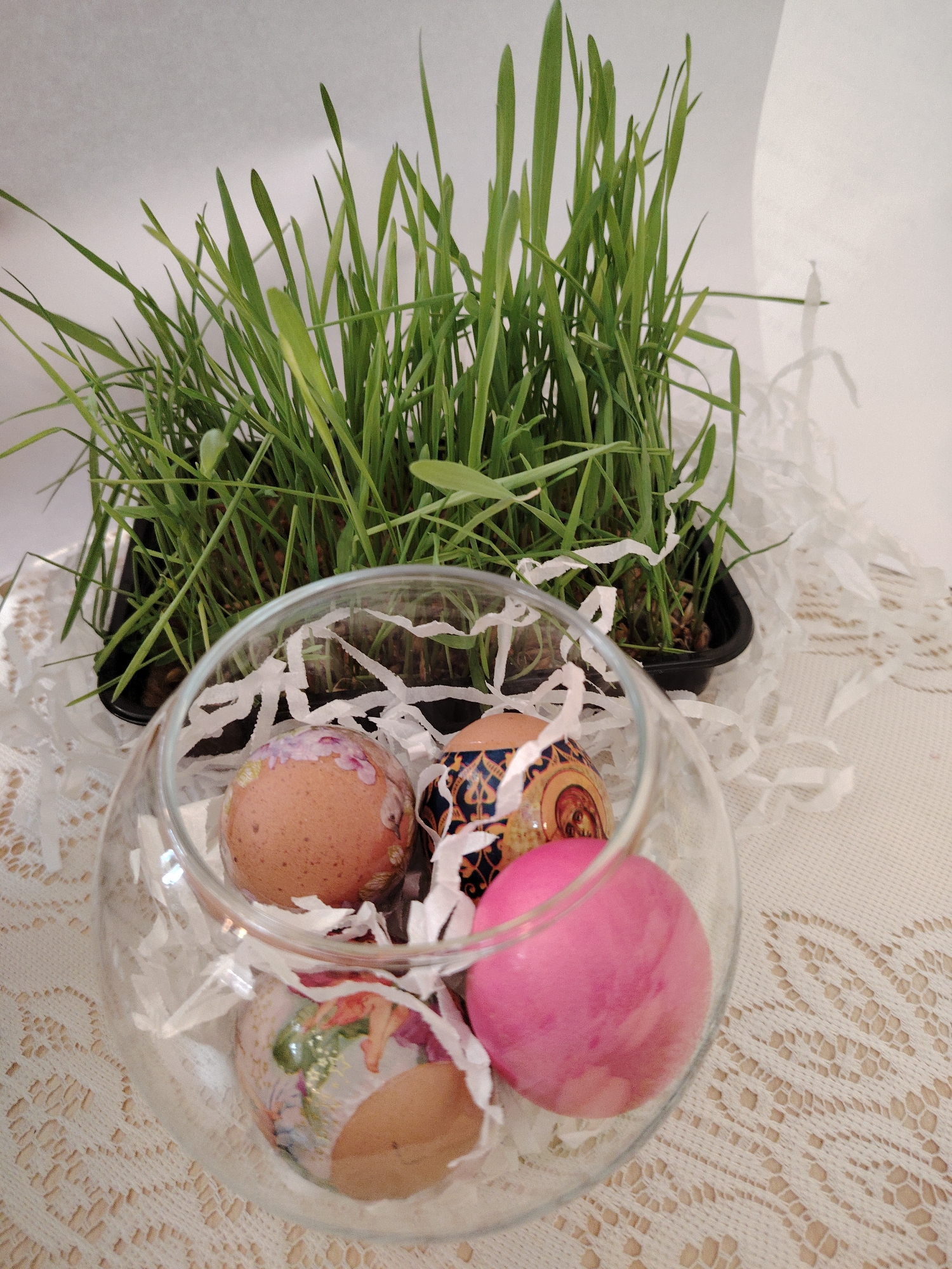 The image shows a still life composition featuring Easter decorations. In the foreground, a clear glass bowl contains four Easter eggs decorated in different styles, surrounded by white shredded paper. Behind the bowl, there is a rectangular black container filled with green grass. The entire arrangement is placed on a beige tablecloth with a lace pattern. The background is a plain white surface. There is no indication of the image being specifically taken in Sarapul, Russia; location information is not provided by the image.