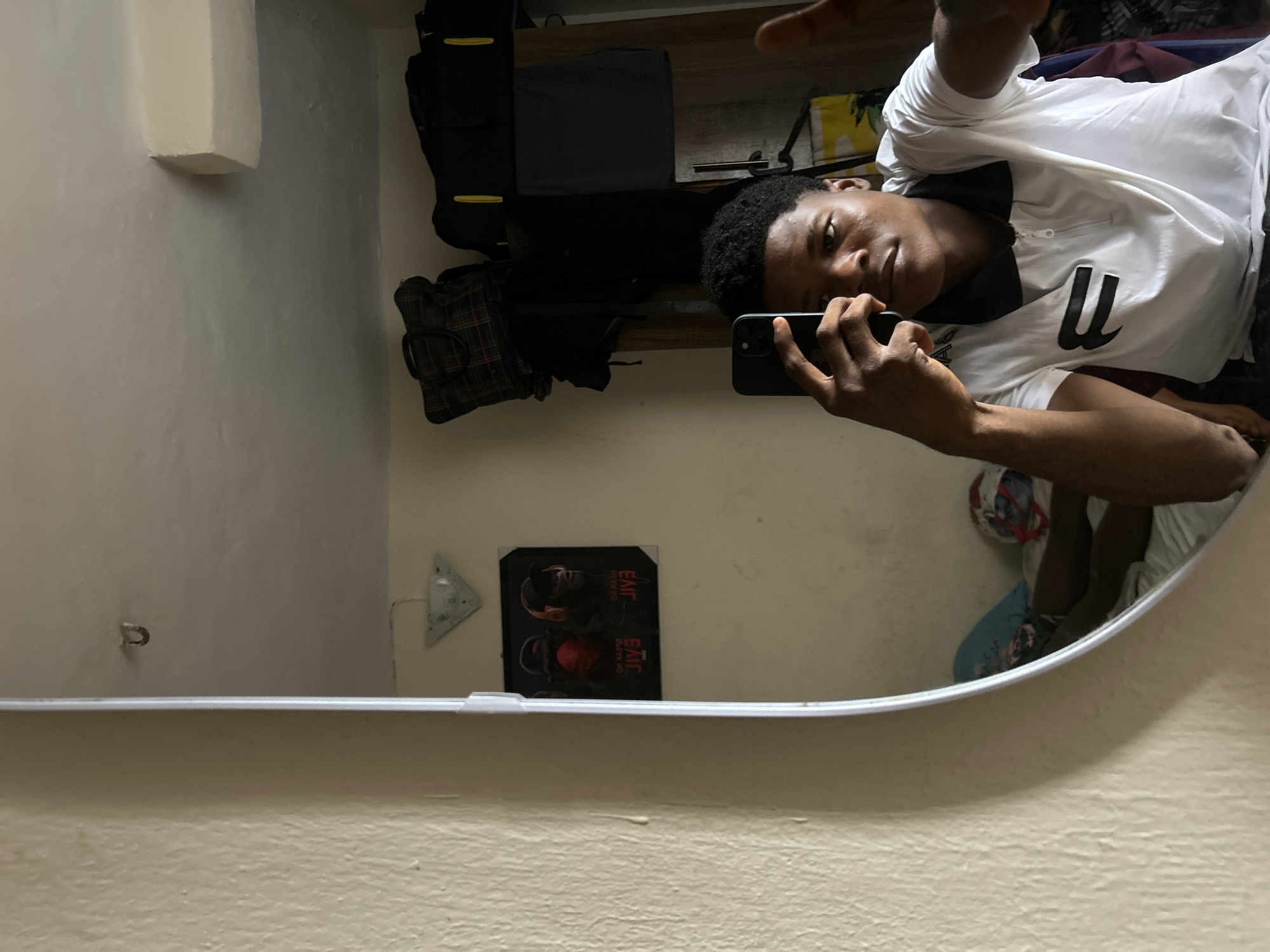 A young adult male is captured in a mirror selfie taken indoors in Ihiagwa, Nigeria. He is positioned in front of a rectangular mirror with a light-colored frame, holding a black smartphone (likely an iPhone with a triple-lens camera array) in his right hand to photograph his reflection. He wears a white polo shirt featuring a black collar, a black zipper placket, and a black stylized letter or symbol resembling an 'F' on the left chest. His left hand grips the phone, while his right hand is partially raised with the thumb visible. He possesses short, dark, curly hair and maintains a neutral expression, looking directly towards the phone's camera.
The room visible in the mirror's reflection appears to be a personal living space. The walls are a pale yellow or beige with a textured finish. On the ceiling, a small, dark, C-shaped hook is visible in the upper left, and a rectangular protrusion is seen in the upper right. To the left of the male's reflection, a triangular wall-mounted light fixture is positioned above a black-framed poster. The poster displays two anthropomorphic figures, one wearing a red face covering and the other headphones, with mirrored text including "JIVA".
Behind the individual, several items are stacked on a brown wooden wardrobe or shelving unit: a dark plaid duffel bag and a larger black bag with yellow accents. Dark clothing items are draped or folded below the bags. The lower portion of the reflection reveals part of a bed with white sheets, a blue pillow, and a white and red object (possibly a helmet or cap) resting on it. The male's lower legs are partially visible, extended on the bed. The room is illuminated by ambient, natural light.