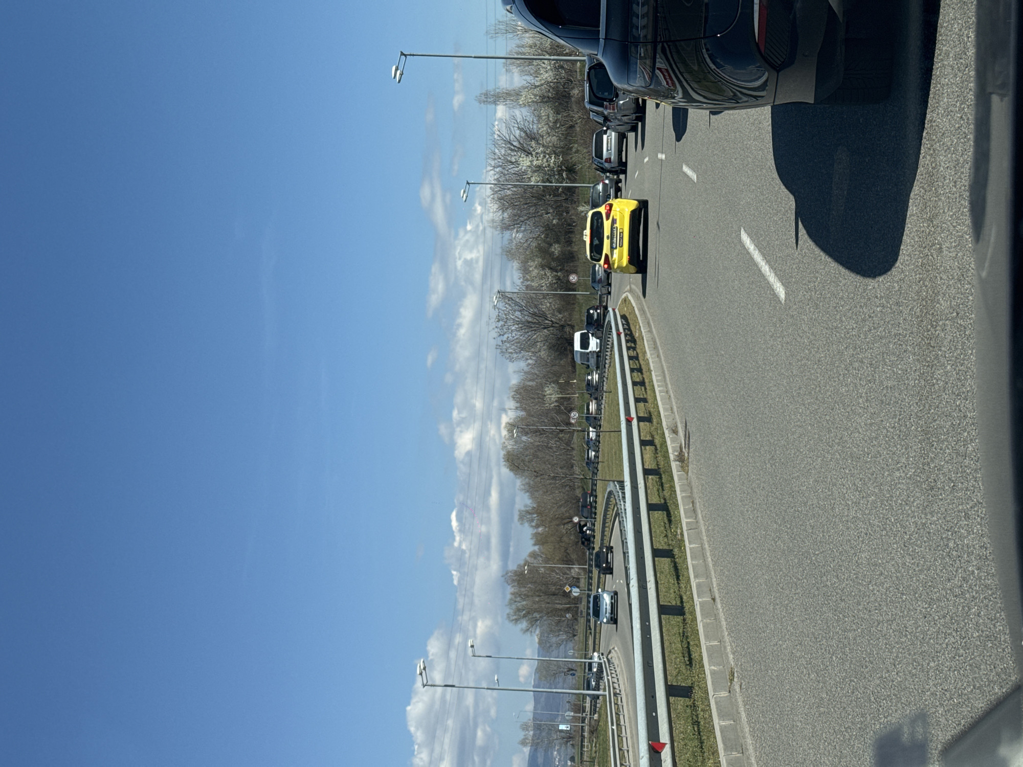 The image shows a traffic jam on a road in Busmantsi, Bulgaria. A yellow taxi is visible in the line of cars. The sky is blue with scattered clouds. Streetlights line the road. Trees are visible in the background.