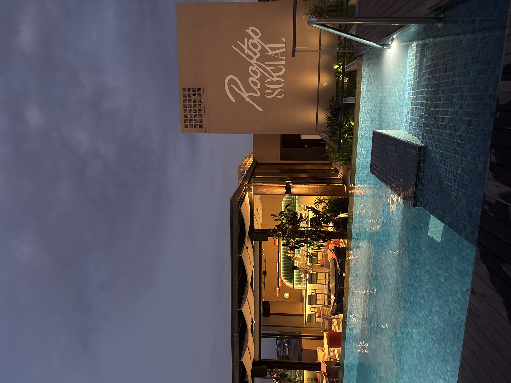 The image depicts a rooftop scene at dusk in North Kuta, Indonesia, featuring a swimming pool in the foreground and a bar area and building facade in the background. The pool is illuminated from within and reflects the twilight sky and surrounding lights. A wooden deck borders the pool, with a metal railing and steps leading into the water.
In the midground, a covered open-air bar area is visible, with several patrons seated at the bar and at tables. The bar is brightly lit, with green tiled accents and warm yellow lighting. Behind the bar, two individuals appear to be working. Lounge chairs and tables with orange cushions are arranged in the seating area.
To the right of the bar area, a building facade is illuminated, displaying the words "Rooftop SOCIAL" in white script. A patterned ventilation grate is visible above the text. Lush green plants are scattered throughout the scene, adding to the ambiance. The overall impression is of a modern and inviting rooftop establishment at the end of the day.