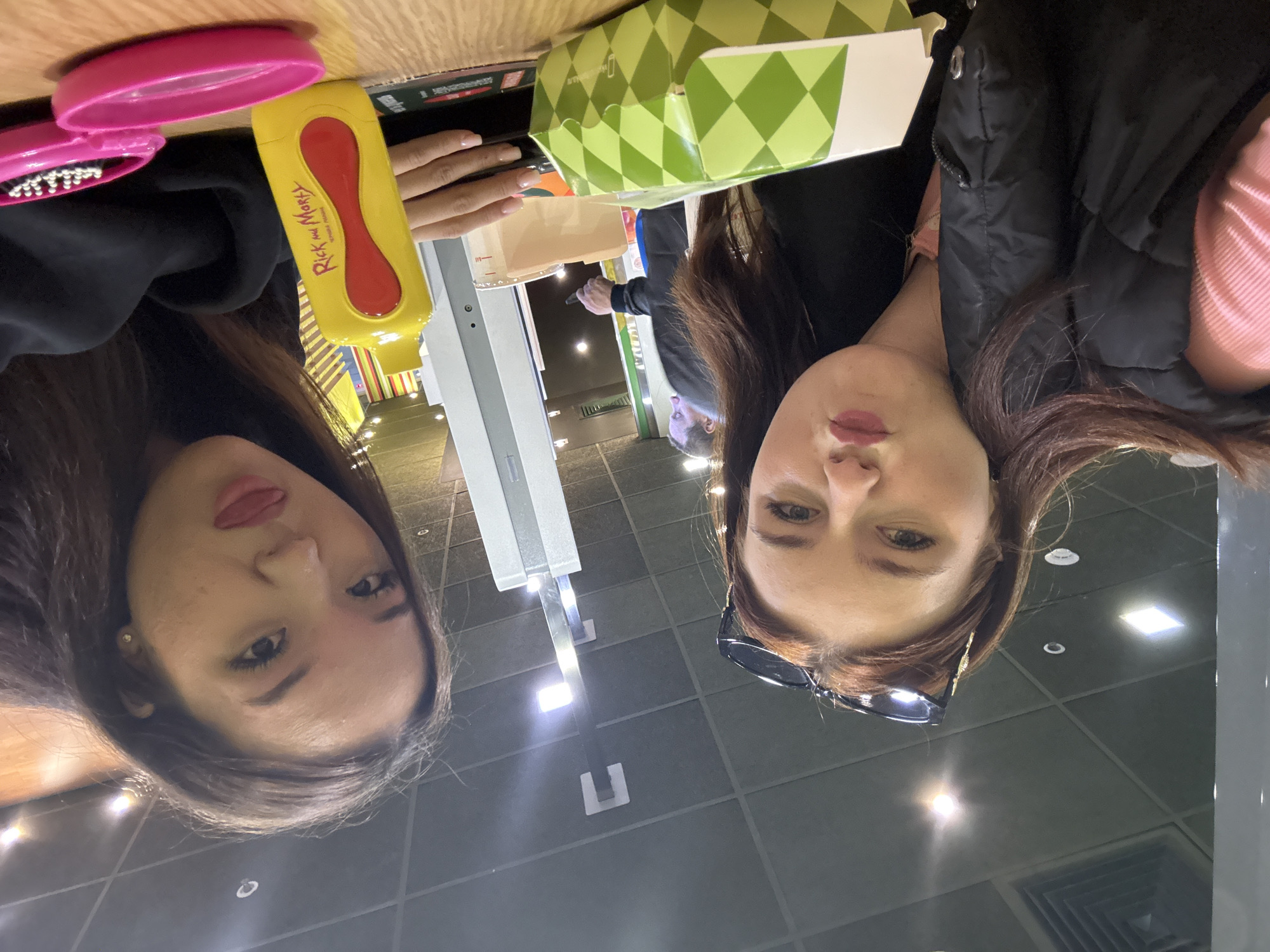 Two women are present in an interior space. One woman is holding a yellow Rick and Morty-branded sauce container and a pink toy. A green patterned box is on the table. The location appears to be a public space with lighting fixtures on the ceiling. The city of Sinyavino, Russia, cannot be confirmed based on the provided image.