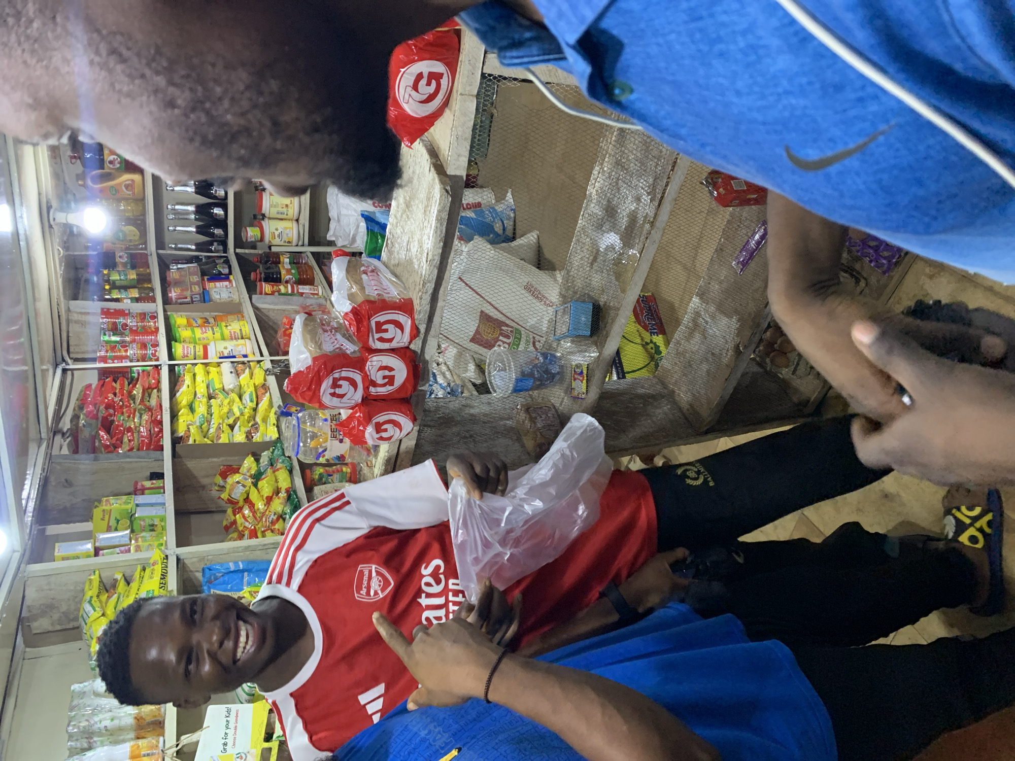 Here's a detailed description of the image:
**Scene:** Interior of a small retail shop in Kabong, Nigeria.
**People:** There are at least three individuals present.
* One is a young Black man smiling and wearing a red and white Arsenal football jersey.
* Another person, is wearing a blue shirt with a Nike swoosh.
* There is a third person in the image.
**Objects:**
* **Goods:** The shop is stocked with a variety of items:
* Packaged food items.
* Bottled beverages.
* Bags of goods.
* **Shop Fixtures:**
* Shelving units hold the merchandise.
* A mesh-covered area, likely for storage.
* A plastic bag.
* Clothes with Nike Logo.
**Actions/Interactions:**
* The man in the Arsenal jersey is smiling. The action is unclear; the individuals may be engaged in a transaction or casual interaction.
**Environment:**
* The shop is well-lit.
* The setting suggests a modest, local business in a residential neighborhood.