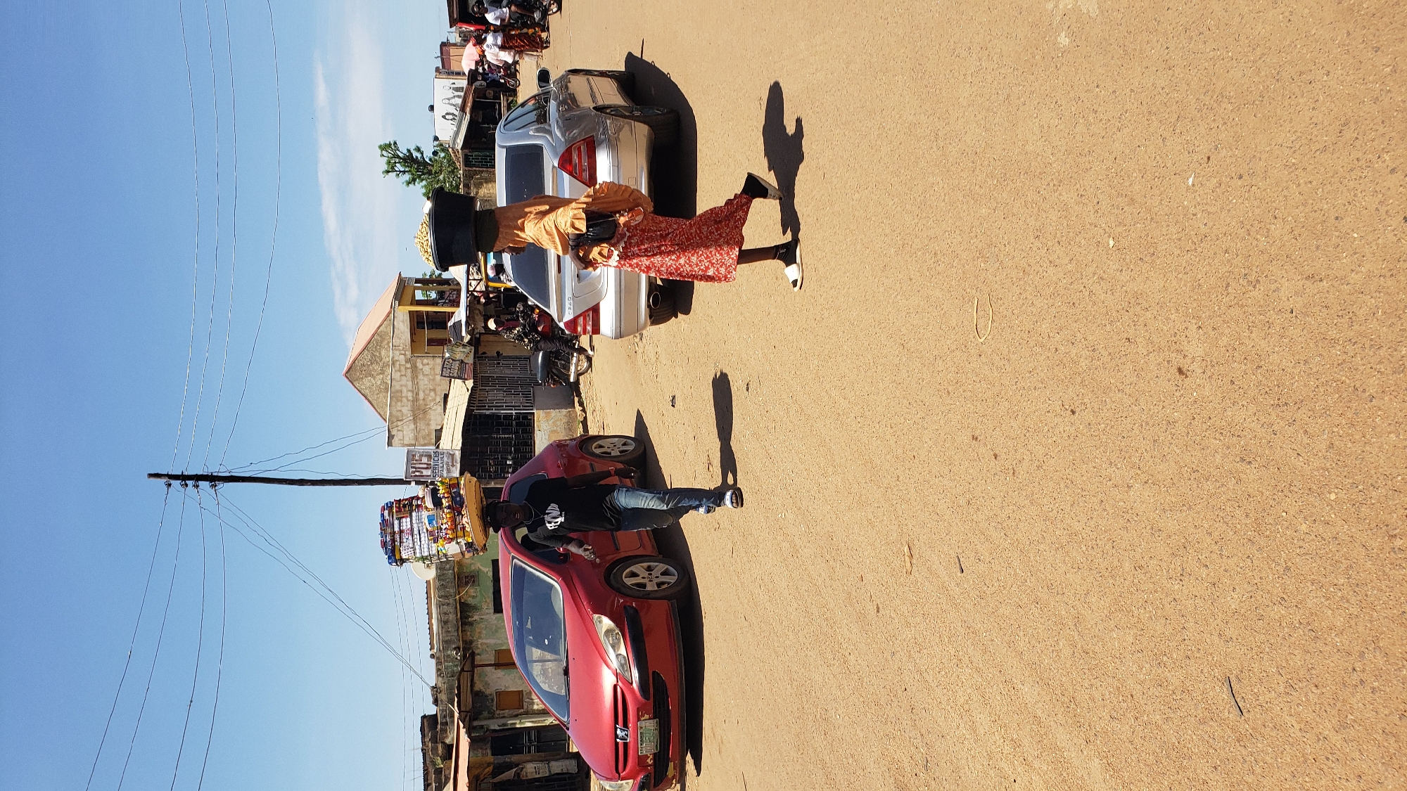 This image depicts a bustling street scene in Sabon Tasha, Nigeria, under a clear, bright blue sky with strong sunlight casting distinct shadows. The unpaved, light brown-orange road occupies the majority of the foreground.
In the left mid-ground, a young man walks forward, wearing a black t-shirt, blue jeans, and light-colored sandals. He carries a large, rectangular tray stacked high with various packaged goods, possibly snacks or provisions, balanced on his head. To his left, a red Peugeot 307 sedan is parked facing left, partially visible.
In the right mid-ground, a woman walks forward, wearing an orange top that covers her head and shoulders, a red and white patterned long skirt, and white sandals with black socks. She carries a large, black round basin, filled with light-colored items (possibly produce), balanced on her head, obscuring her face. A small, dark-colored shoulder bag hangs from her right shoulder. Behind her, a silver Mercedes-Benz C-Class sedan is parked or moving slowly, facing right.
The background features various single-story and two-story buildings, likely shops or residences, lining both sides of the street. A prominent feature is a tall, dark wooden utility pole with multiple power lines extending across the sky, located between the two main figures. A white sign with red lettering reading "POS SERVICES AVAILABLE HERE" is visible behind the red car. In the far background on the right, more people are visible, some on motorcycles, and one person holding an umbrella, suggesting further activity down the street. Small pieces of debris, including dark, thin objects and a light-colored U-shaped object, are scattered on the dusty road surface.