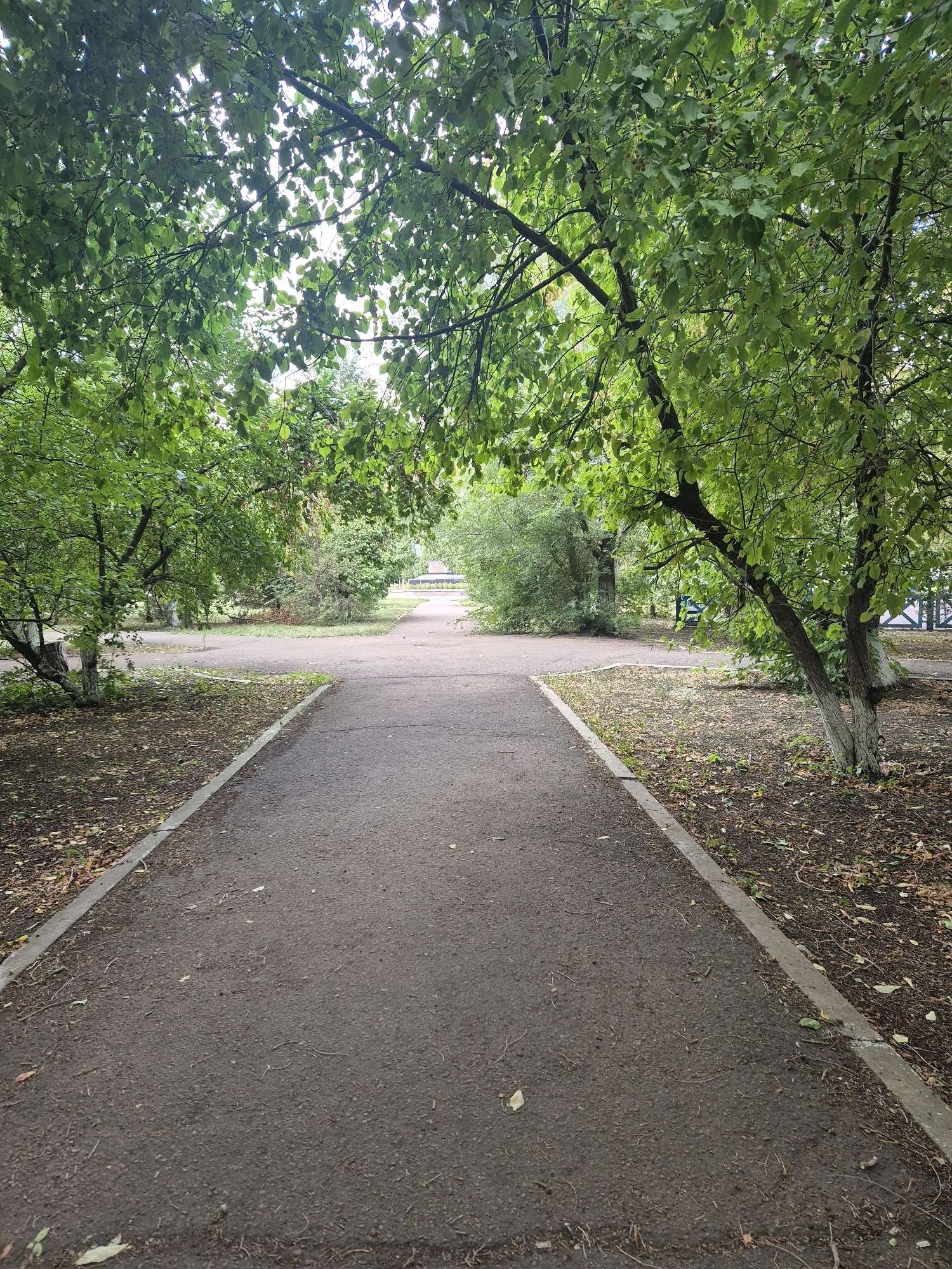 The image shows a paved pathway in a park-like setting. The pathway splits and rejoins in the foreground, with grass and fallen leaves on either side. Mature trees with green foliage overhang the pathway, casting dappled shade. In the background, more trees and bushes are visible, along with a glimpse of a blue and white fence. The sky is overcast, suggesting a cloudy day. The location is the park located at the intersection of Abylay Khan Avenue and Bukeykhan Street in Kokshetau, Kazakhstan.