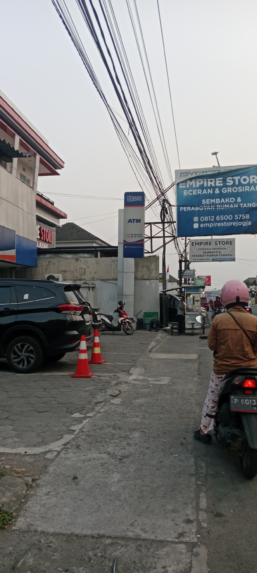 The image captures a street scene in Maguwoharjo, Indonesia. On the left, a black SUV is parked, with two orange traffic cones in front of it. Adjacent to the SUV is a building with red trim and signage that includes the text "E STORE" and "LAYAN". To the right of the building, a blue sign for "BRI ATM" is visible. Further to the right, a large blue banner for "EMPIRE STORE ECERAN & GROSIR SEMBAKO & PERABOT RUMAH TANGGA" hangs overhead, along with a smaller, similar banner below it. A person on a motorcycle wearing a pink helmet and patterned pants is in the foreground, facing away from the camera, with the rear of their motorcycle visible. The license plate on the motorcycle reads "P 6013". In the background, there are other vehicles, structures, and a power pole with numerous cables. The sky is overcast.