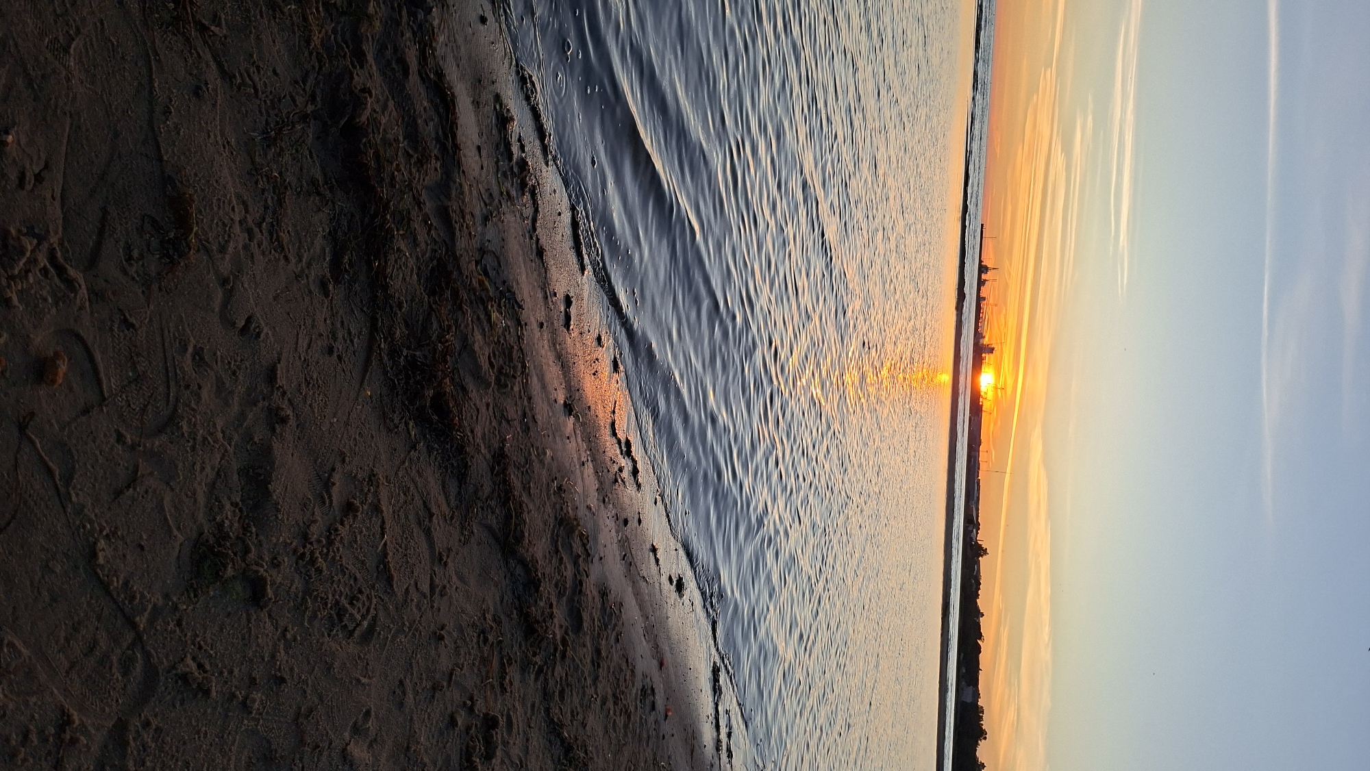 The image captures a sunset over the Baltic Sea in Tallinn, Estonia. The sun is low on the horizon, casting a warm orange glow across the sky and reflecting on the water. Silhouettes of industrial structures, possibly cranes and buildings, are visible in the distance along the coastline, suggesting an industrial or port area. The foreground shows a sandy beach with some debris and subtle footprints. The water is calm with gentle ripples, and the sky transitions from a pale blue at the top to shades of orange and yellow near the horizon, with thin wispy clouds.