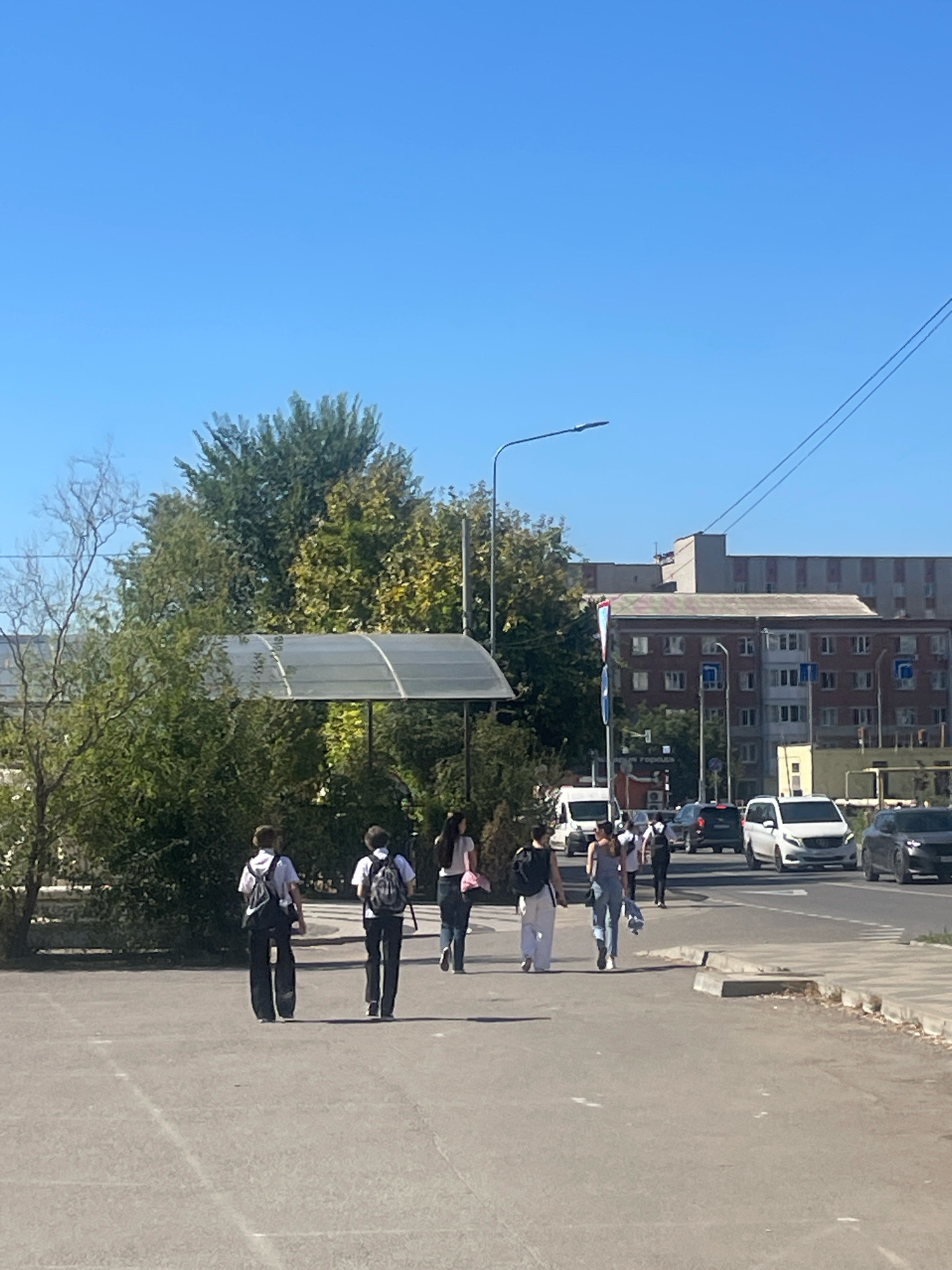 In this outdoor, daytime scene, a clear blue sky stretches above a street in the city of Krasnodar, Russia. On the left side of the image, two young males, presumably students, walk away from the camera, each wearing a backpack. One is dressed in a white shirt and dark pants, the other in a black shirt and dark pants. To their right, a group of young women also walks away from the camera. The foreground consists of a paved area, likely a sidewalk or pedestrian walkway, transitioning to a street with vehicular traffic in the midground. Several vehicles, including a white van and a white SUV, are visible on the street. Tall trees and lush green foliage are present on the left, partially obscuring a covered structure with a translucent roof, possibly a bus stop or a covered walkway. In the background, multi-story apartment buildings with red brick facades and windows are visible, along with a taller, grey building. A street lamp stands on the right side of the street. The overall impression is of an ordinary day in an urban environment, with people moving about their daily routines.