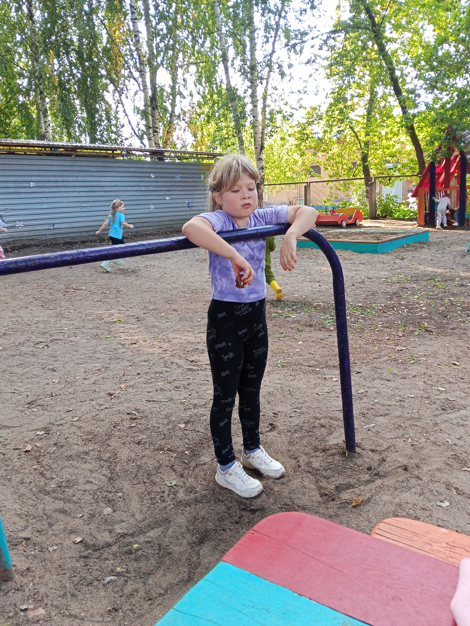 In Chaikovsky, Russia, a young blonde girl, wearing a purple t-shirt and black leggings with white lettering, stands in a sandy playground. She is leaning on a purple metal bar. Her eyes are partially closed, and she appears to be resting. In the background, another young girl in a blue shirt runs. A playground structure with red and blue elements is visible further in the background, along with trees and a grey metal fence. The ground is covered in sand and scattered leaves.