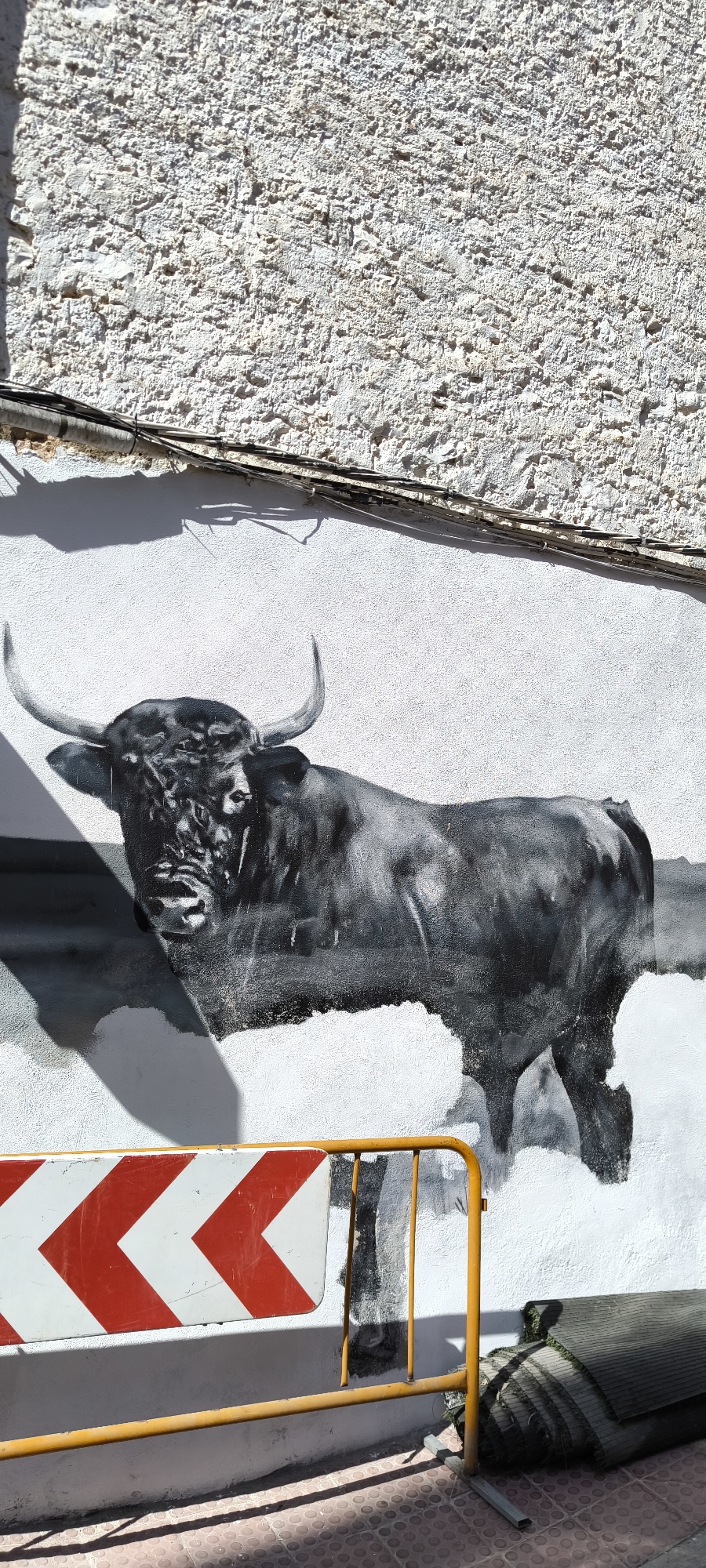 A black and white mural of a bull adorns a white textured wall. The bull is depicted from the chest up, facing forward with its head turned slightly to the left. Its horns are prominently displayed. The mural is rendered with a gritty, almost photographic style, with heavy shadows and highlights emphasizing the bull's musculature and facial features. In the foreground, a yellow metal barrier with red and white chevron markers partially obscures the lower portion of the mural. To the right of the barrier, a rolled-up dark green object, possibly artificial turf, is leaning against the wall. The ground in front of the barrier is paved with red and grey tiles, casting shadows from the barrier and an unseen object. Electrical cables are visible running horizontally across the upper part of the wall, casting a thin, elongated shadow. The overall scene suggests an urban setting, possibly a street or walkway in Torreblanca, Spain, where art and temporary traffic control measures coexist. There are no people visible in the image.