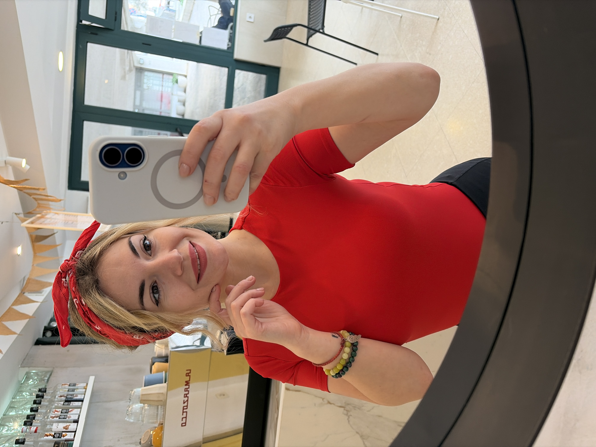 A fair-skinned woman with blond hair wearing a red bandana and red shirt, and a beaded bracelet on her left wrist is taking a selfie in a mirror. The phone is light grey and has two camera lenses. In the background, there is a shop with glass shelves holding bottles and jars. A coffee machine is visible. Outside the shop, a black chair and a window offer a glimpse of the outdoors. The location is likely in Budva, Montenegro, given the mention of it in the instructions.