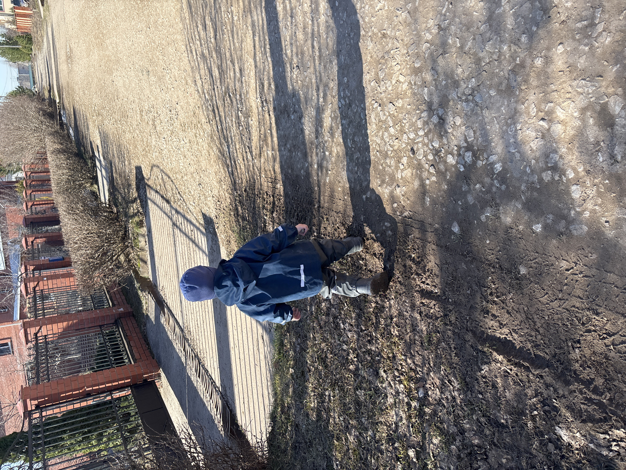 A child in a blue coat and hat walks on a dirt path in Ферма, Russia. The path is edged by a metal fence and a row of bushes on one side, and a gravel road with tree shadows on the other. The ground is a mix of dirt, gravel, and some patches of grass and dead leaves. The weather appears sunny.