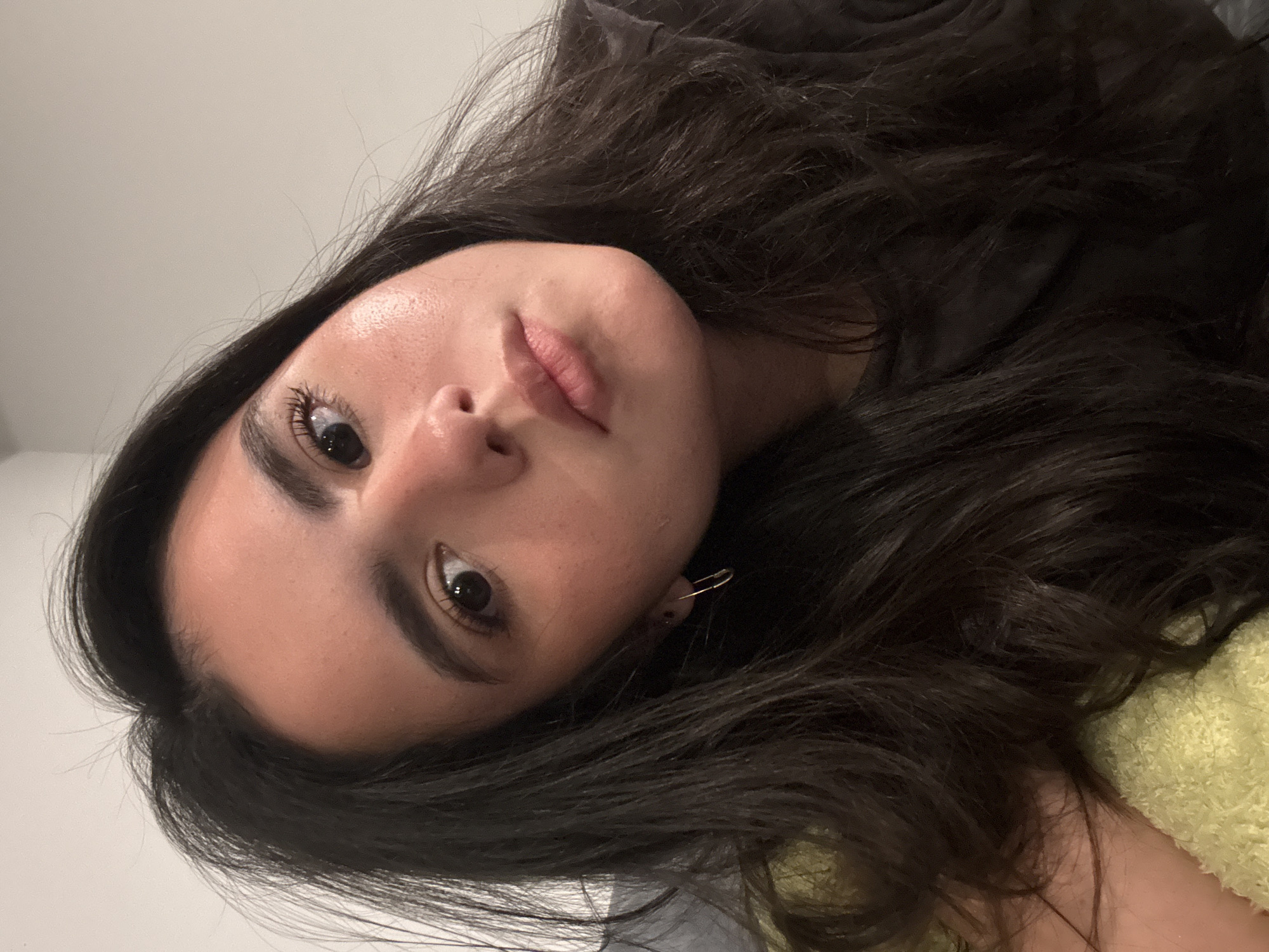 A close-up portrait of a woman with long, dark, wavy hair. Her face is angled towards the camera, showcasing her facial features. She has dark eyes, full lips, and defined eyebrows. She is wearing a dark-colored shirt. A safety pin is visible in her ear. The background appears to be a light-colored wall. There is no information present to suggest that the photo was taken in Austin, United States of America.