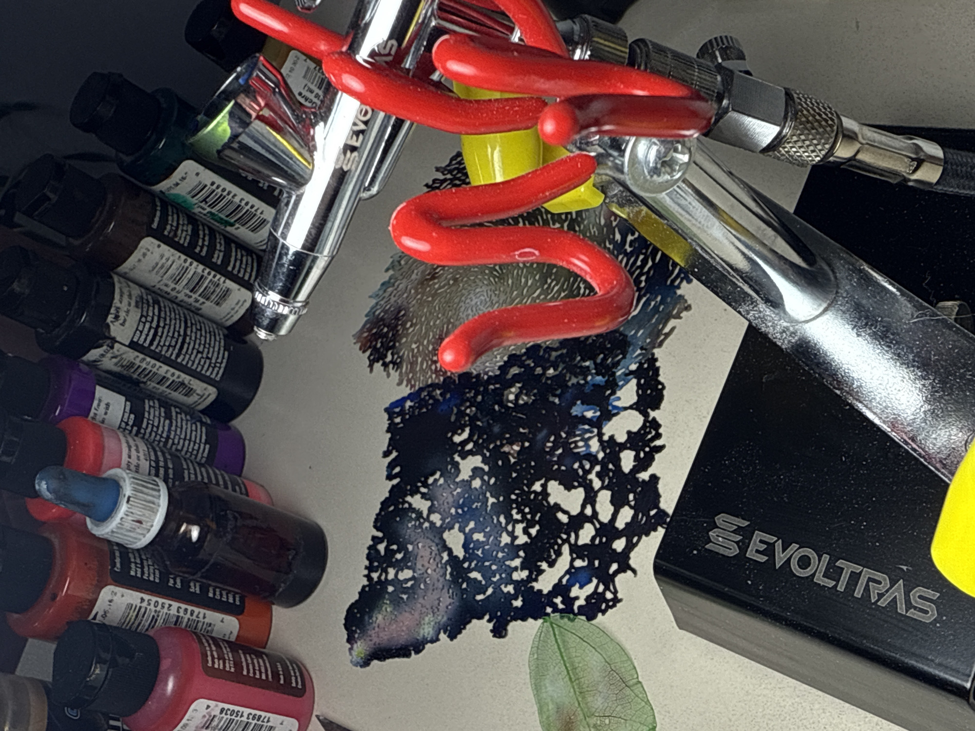This image shows a collection of art supplies. There are multiple small bottles of acrylic paint, with colors ranging from greens and browns to purples, oranges, and reds. An airbrush is positioned among the paint bottles, with a red airbrush hose attached. There's also a small leaf on the surface. The lid of an "EVOLTRAS" box is visible on the right. There is a black ink blot in the center of the scene. The labels on the paint bottles have text, including the numbers "7 17893 15038", "17893 25054", and "7 17893 20002". The lid says "Ochre 10 ml."