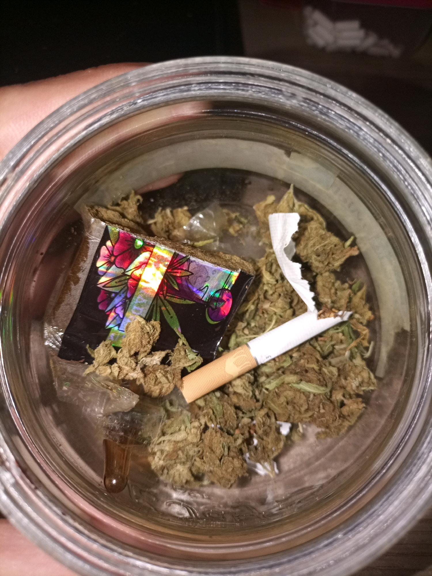 A glass jar contains dried cannabis buds, a cigarette, and a small piece of holographic material with a floral design. The jar is held by a hand, partially visible at the top left of the image. The scene is an indoor, close-up shot, with no discernible environmental context suggestive of Tarragona, Spain.