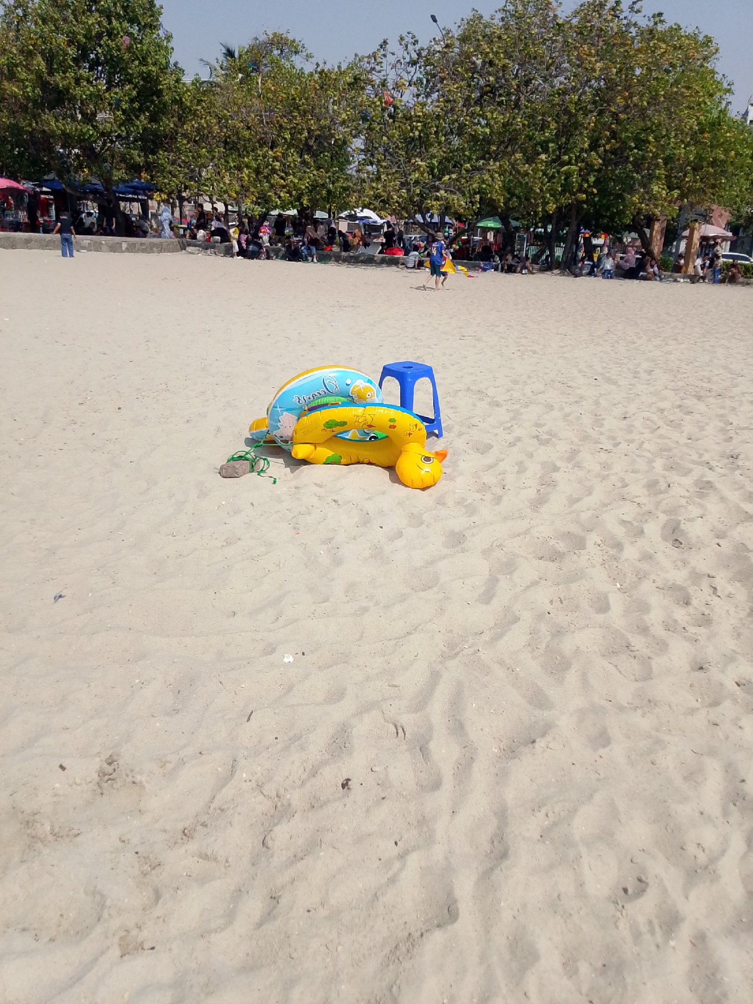 The image depicts a sandy beach with several inflatable toys in the foreground. Specifically, a yellow turtle-shaped inflatable is prominently displayed, partially deflated and lying on its side. Next to it, a blue plastic stool is visible. In the background, a line of trees with dense foliage forms a backdrop to a crowd of people seated and standing under makeshift shelters, likely vendors and beachgoers. The scene suggests a public beach environment, potentially Ancol Beach in North Jakarta, Indonesia, known for its recreational activities and street vendors. The sand is light-colored and appears somewhat dry, with subtle footprints and ripples indicating recent activity. The sky is clear and bright, suggesting a sunny day.