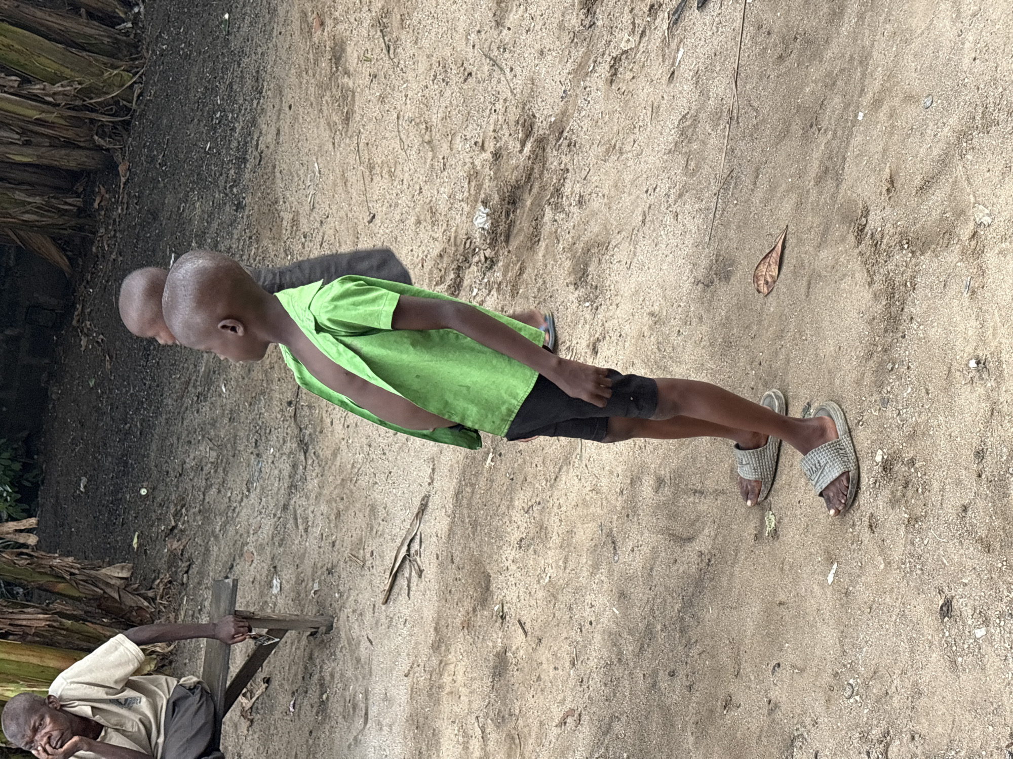 The scene is in Ikot Ekpene, Nigeria. An elderly man is seated, appearing worn. He rests his head in his hand, leaning against a rudimentary wooden structure. Nearby stand two young boys. The child closest to the camera wears a green shirt and black shorts, while the other's attire is obscured. The ground is sandy and uneven.