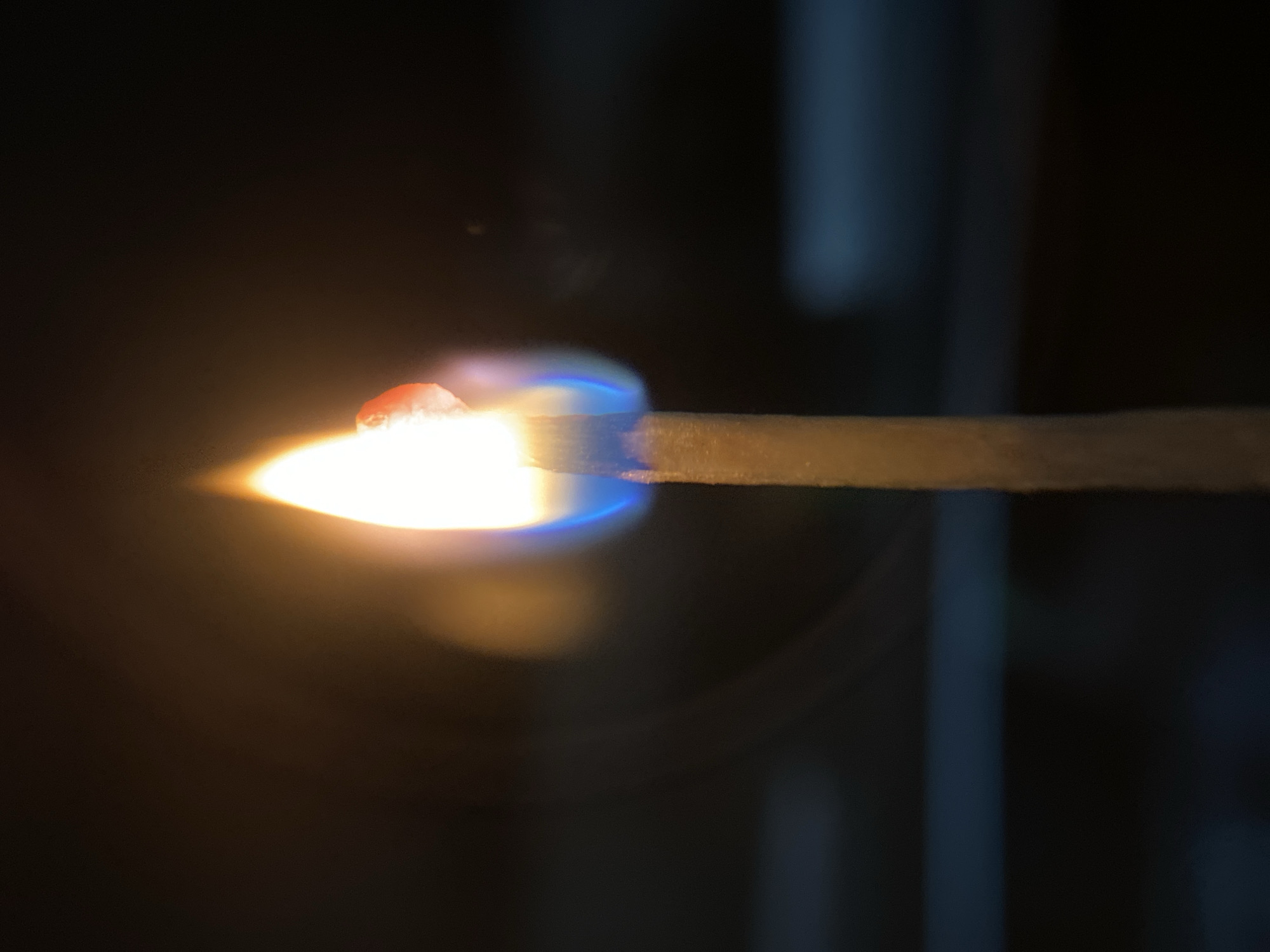 A close-up, vertical, macro photograph focuses on the burning tip of a single wooden matchstick. The flame is a bright white and yellow, with a distinct blue hue at its base where it connects to the match. A small, reddish-orange globule, possibly molten material from the match head, is visible on the right side of the flame. The matchstick itself appears to be made of wood, with a rough texture and a light brown color. The background is dark and out of focus, with horizontal lines of muted blue and black suggesting an indoor environment, possibly a window or blinds. The lighting is primarily from the match flame, casting a warm glow on the immediate surroundings. There are no people or other living beings present in the image.