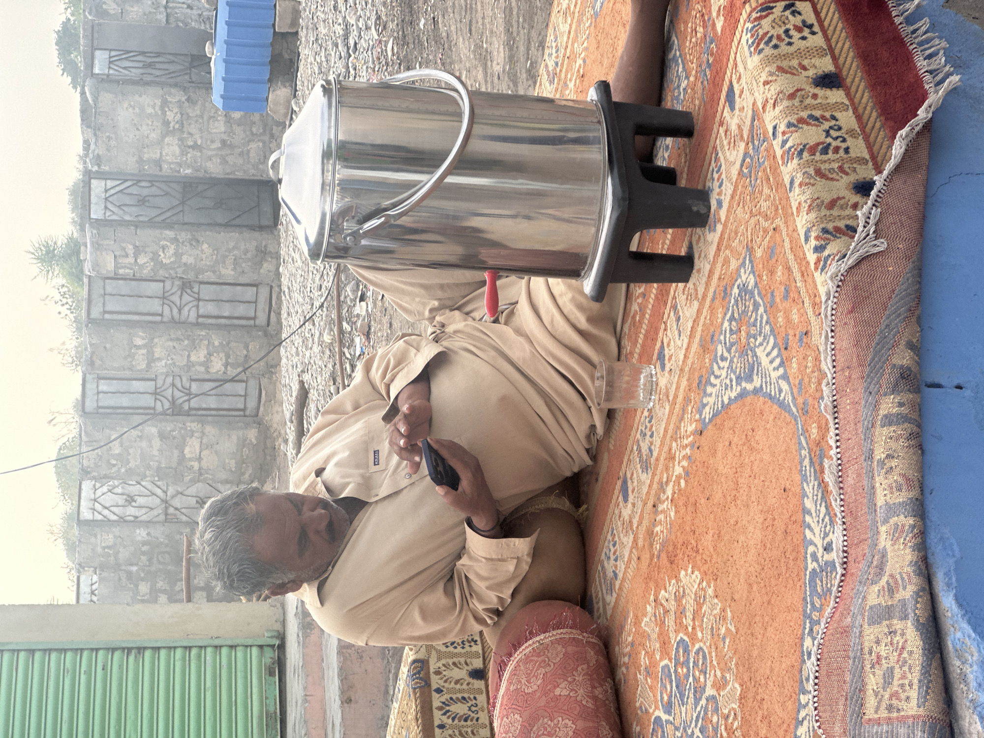 A middle-aged man with gray hair is sitting on an ornate rug in Unknown, Unknown. He is holding a black smartphone. A large stainless steel water cooler sits next to him on a black plastic stand, with a drinking glass nearby. The environment includes a building with barred windows and a green corrugated metal gate. A blue container sits on the roof of the building. The area appears slightly hazy.