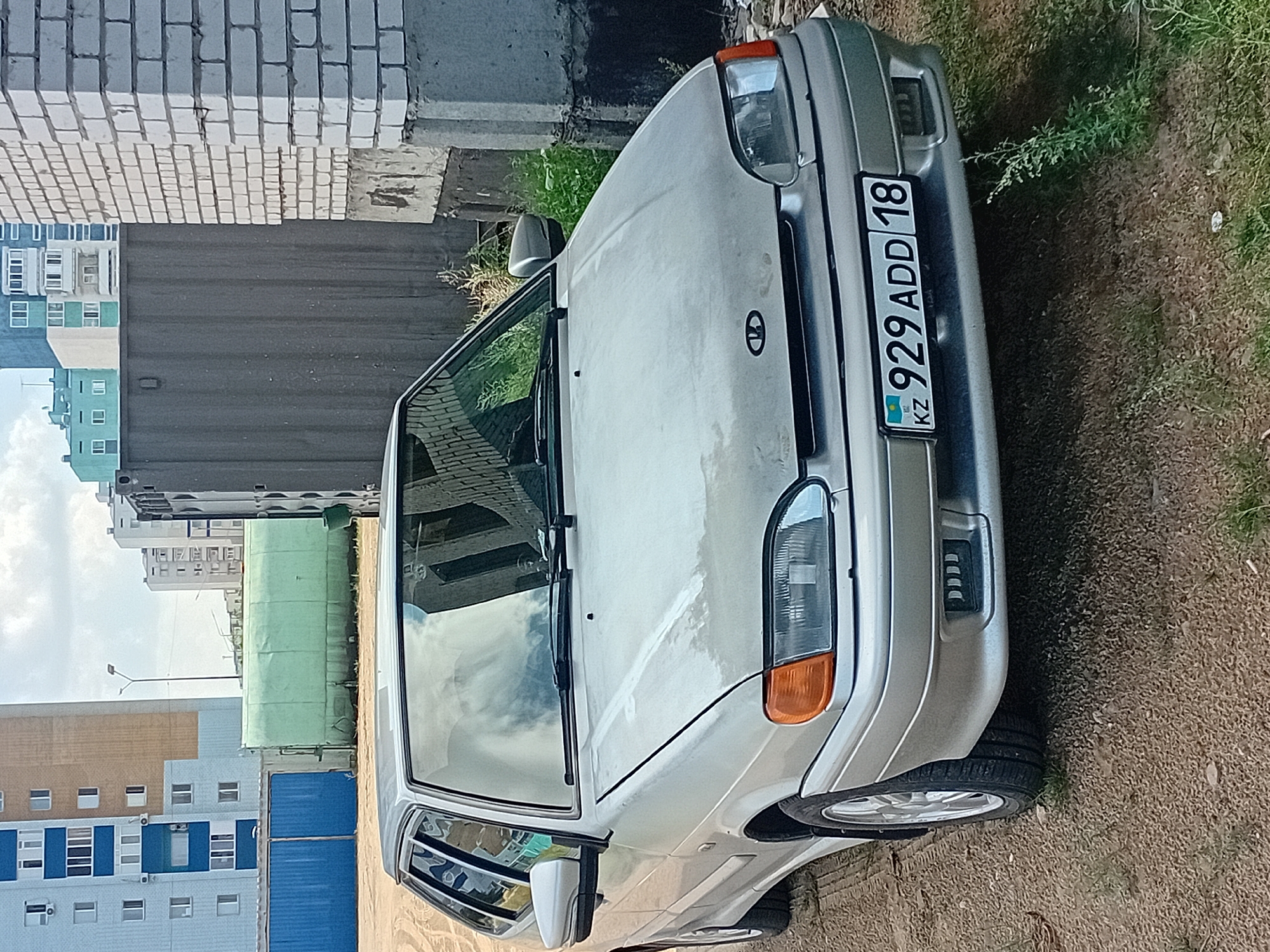 A silver sedan, license plate "929 ADD 18", is parked on a gravel lot in front of a brick building. The car is angled to the left, with its front facing the viewer. In the background, there are several multi-story buildings and a large, greenish structure. The sky is cloudy. The scene is likely located in Semey, Kazakhstan, given the license plate.