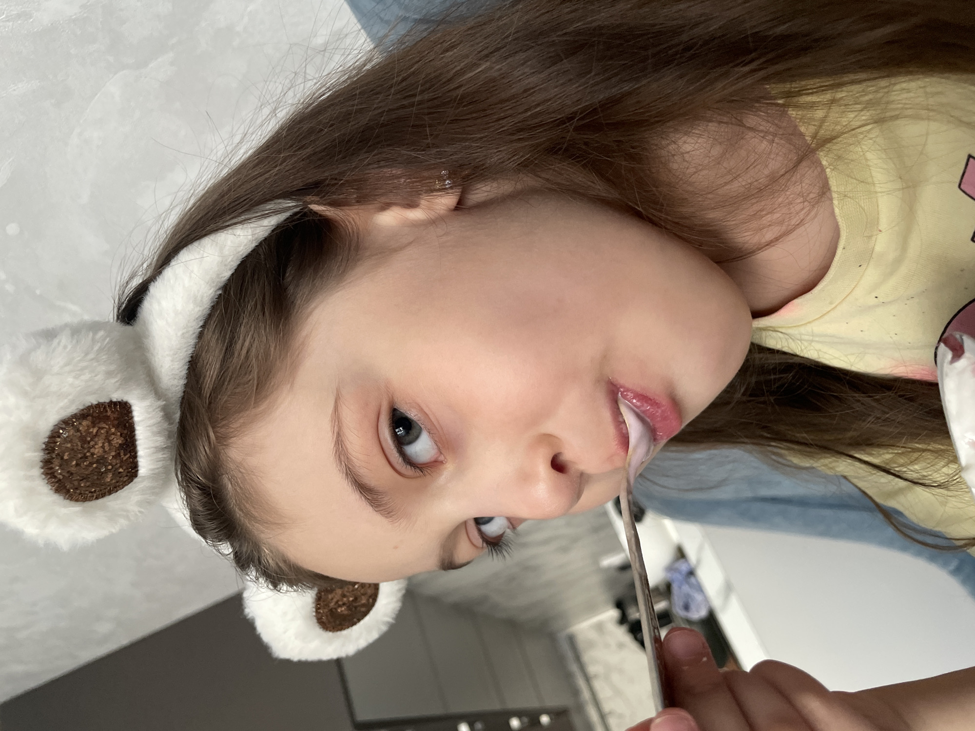 A candid, close-up portrait captures a young girl with long, straight brown hair, positioned on her side. She is wearing a whimsical white, furry headband adorned with two ear-like attachments, each featuring a shimmering brown, glittery circular center, resembling animal ears (possibly bear or panda). Her light-colored eyes are wide open, looking directly at the viewer with a neutral and engaging expression.
She wears a light yellow t-shirt and another light blue garment is visible behind her shoulder. A metallic utensil, appearing to be a spoon or spatula, is held in her hand, reaching towards her mouth. A white, creamy substance, likely yogurt or a similar snack, is visible on her lower lip and appears to be dripping from her mouth, indicating she is actively eating.
The background is softly blurred, revealing an indoor setting with light-colored walls and hints of darker elements, suggesting a home environment such as a kitchen or dining area. The lighting is bright and even, possibly natural daylight or well-diffused indoor lighting. There is no visible text in the image. The scene conveys a simple, everyday moment of a child enjoying food, set in Izhevsk, Russia.