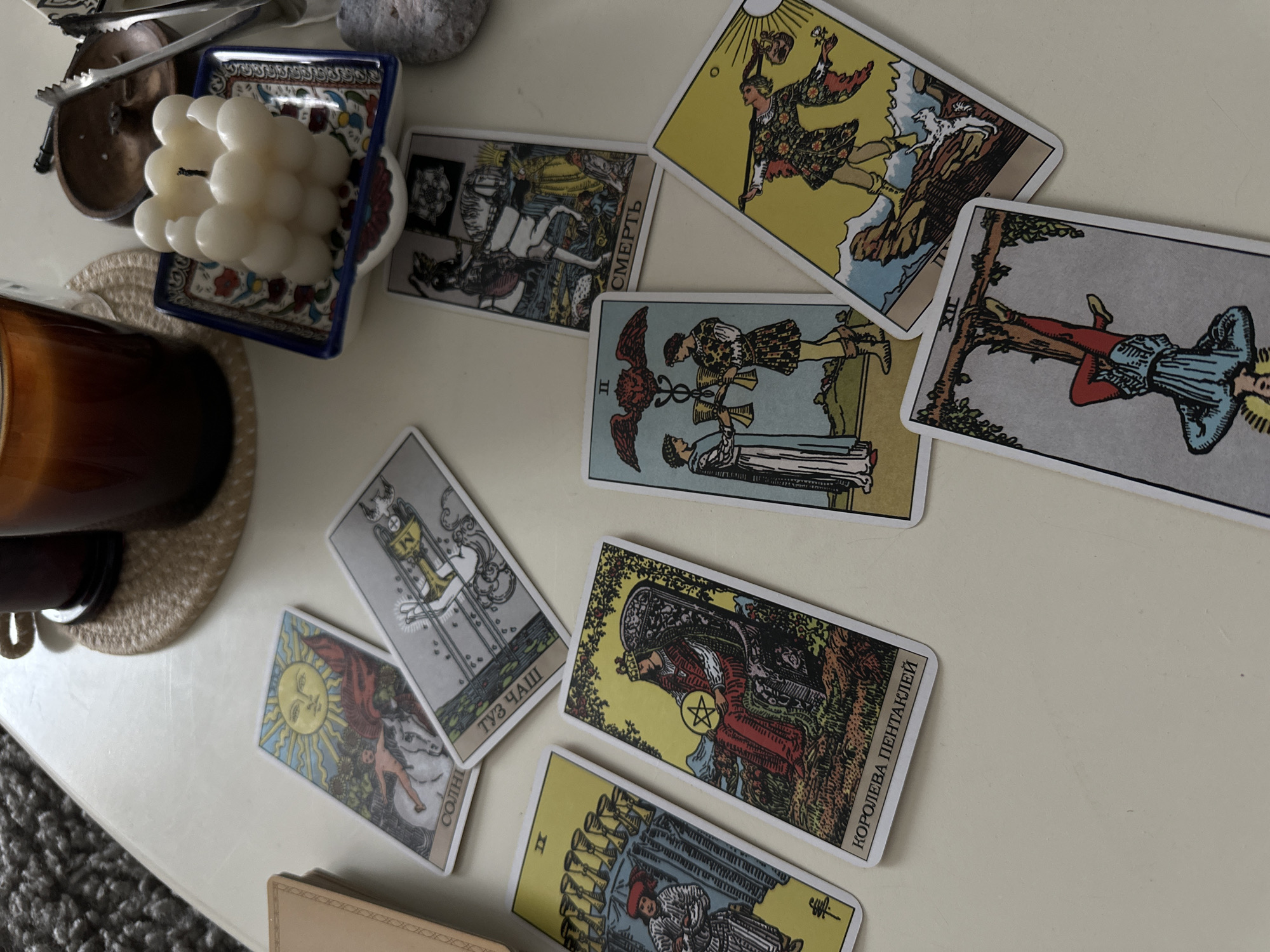 This overhead shot captures a collection of objects arranged on a light-colored surface, suggesting an indoor setting. The primary focus is a spread of numerous tarot cards, indicative of a tarot reading session.
Among the cards, several are clearly identifiable: The Fool is visible in the top right, depicting a figure and a dog by a cliff; The Hanged Man (XII), inverted, shows a figure suspended by one foot; The Death card, inverted, features a skeleton on horseback with the Cyrillic word "СМЕРТЬ" (Death); The Two of Cups (II) displays two figures exchanging goblets; The Sun card, inverted, portrays a child on a white horse under a radiant sun with "СОЛНЦЕ" (Sun) inscribed; The Ace of Cups, partially visible, has "ТУЗ ЧАШ" (Ace of Cups); The Queen of Pentacles, partially visible, shows a regal figure holding a pentacle and labeled "КОРОЛЕВА ПЕНТАКЛЕЙ" (Queen of Pentacles); and a portion of the Ten of Cups (X), inverted. The visible text on the cards is in Cyrillic, suggesting a Russian or Ukrainian language deck.
Adjacent to the cards, a white, multi-lobed candle rests in a small, square ceramic dish adorned with blue trim and a vibrant floral pattern. To its left, a dark amber-colored glass jar, likely a candle or diffuser, sits on a woven, circular coaster. Other background elements include a small wooden bowl, silver tongs, a smooth grey stone, and a glimpse of a textured dark grey rug or blanket in the lower left corner. The lighting is soft and ambient, providing no specific cues for the time of day. The arrangement of items points to a moment of contemplation or spiritual practice, possibly in a personal space.