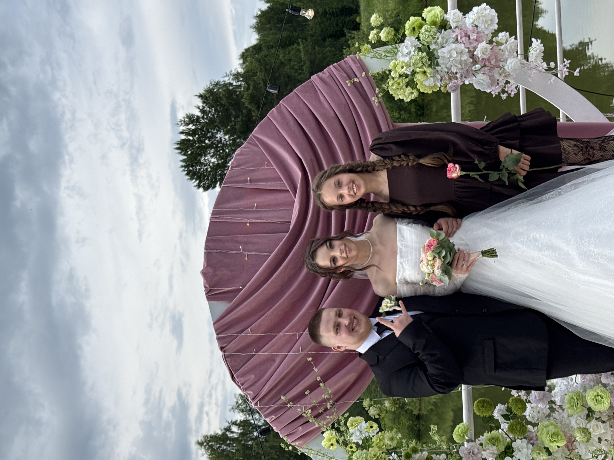 This media file shows three people at an outdoor wedding ceremony in Votkinsk, Russia. The subjects are a bride in a white wedding dress, a groom in a black suit, and a young girl, likely a bridesmaid or flower girl, in a dark brown dress. The bride and groom are standing together, with the bride holding a bouquet of pink and white flowers. The young girl is standing beside them, her hair in braids, and she is smiling at the camera.
The scene is set against a backdrop of a large, mauve-colored fan-shaped curtain, adorned with fairy lights. In the background, there are trees, a body of water, and a cloudy sky, suggesting the event is taking place during the day. Floral arrangements of white and green flowers are visible, adding to the celebratory atmosphere. The overall mood appears joyful and festive. There is no visible text in the media file.