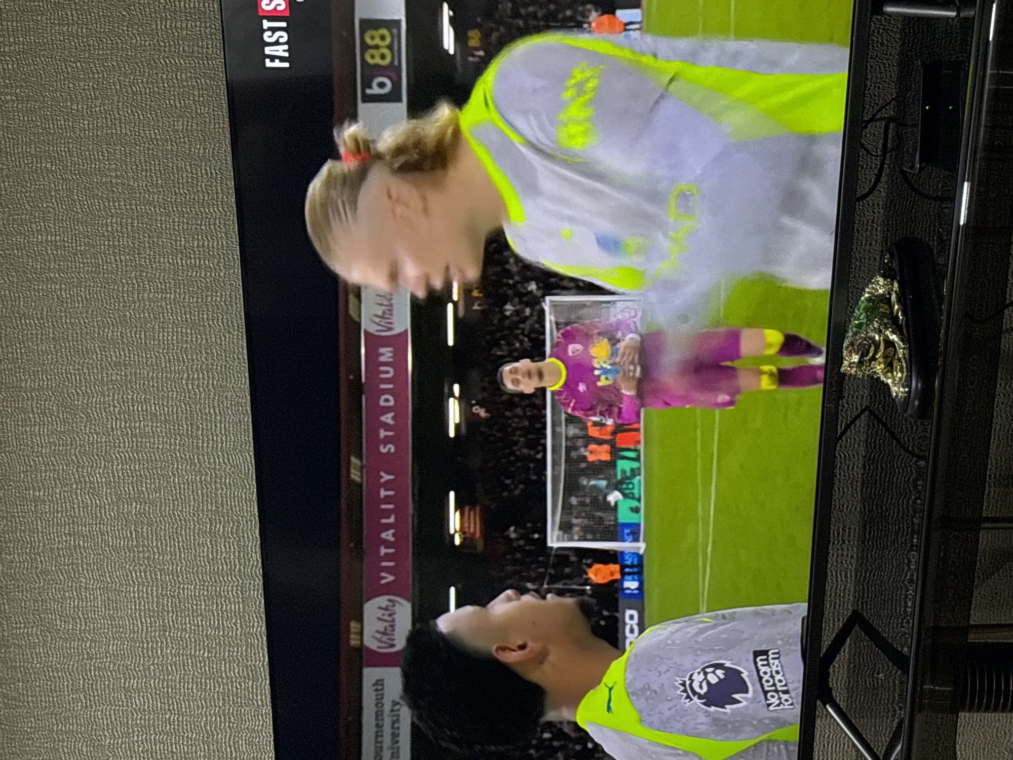 The image shows a television screen displaying a soccer match. In the foreground, two players wearing white jerseys with neon green accents are visible. One player has light skin and blonde hair pulled back in a ponytail, while the other has dark hair. Behind them, the soccer field stretches out with a goalkeeper in a purple jersey near the goal. The stadium is identified as "Vitality Stadium." Text on the screen displays "Bournemouth University" and "Vitality STADIUM." A logo with "No room for racism" is also visible on one of the players' jerseys. "FAST S" and "b 88" are displayed at the top of the screen. There are glimpses of objects on a black table beside the TV. The location of the image is not Yerevan, Armenia.