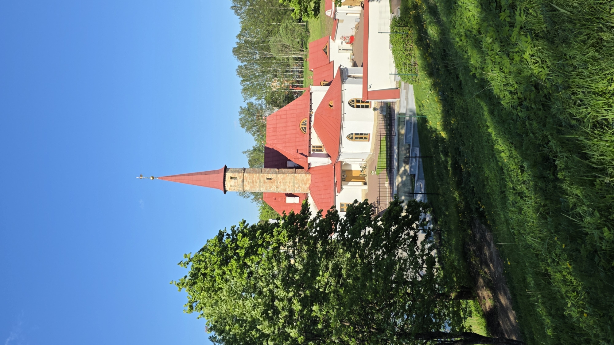 The image shows the Priory Palace located in Gatchina, Russia. The palace has a predominantly white facade with a striking red roof. A tall, slender tower with a pointed spire stands prominently, contributing to the architectural silhouette. Trees and greenery surround the structure, and a grassy area is present in the foreground. The sky is a clear blue, providing a stark contrast to the colors of the palace and the natural surroundings.