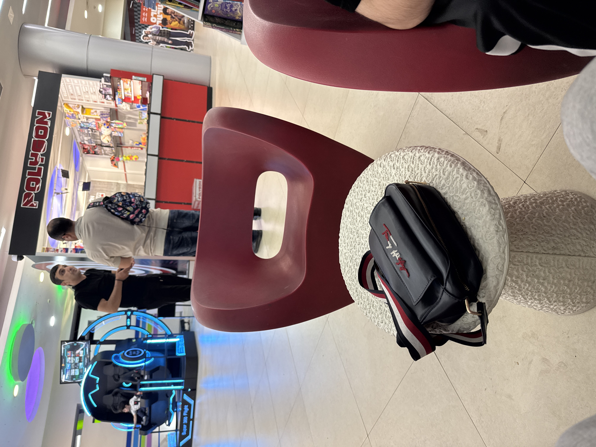 The image captures an indoor scene within what appears to be a modern entertainment or retail complex, likely a shopping mall, in Yerevan, Armenia. The floor is light-colored and tiled, reflecting the bright artificial lighting.
In the immediate foreground, the lower half of a person wearing black shorts with white stripes is visible, seated on a sleek, maroon-red chair. Adjacent to this, a dark blue "TOMMY HILFIGER" crossbody bag, featuring red and white embroidered text and a striped strap (red, white, blue), rests on a textured white cylindrical stool. Another similar maroon-red chair, with a distinctive cutout design, is positioned further back.
Four individuals are visible in the scene. In the midground, a person with dark hair, wearing a light-colored top and jeans, stands with their back partially turned, sporting a colorful patterned fanny pack. They are looking towards a second person, also with dark hair, dressed in a black t-shirt and black pants, who stands next to a "Super 360 Flight" gaming simulator. This simulator, illuminated with bright blue neon-like lights, contains a third person seated inside, actively engaging with the game.
To the left of the scene, a store entrance is prominent, featuring a tall vertical sign with "POLYGON" written in bold red letters on a black background. The store's interior displays various products, suggesting it might be a toy, game, or collectibles shop. In the background, other commercial signage and posters are visible, including one partially obscured sign with the text "ILO". The overall impression is one of a lively public space where people are engaged in leisure and shopping activities.