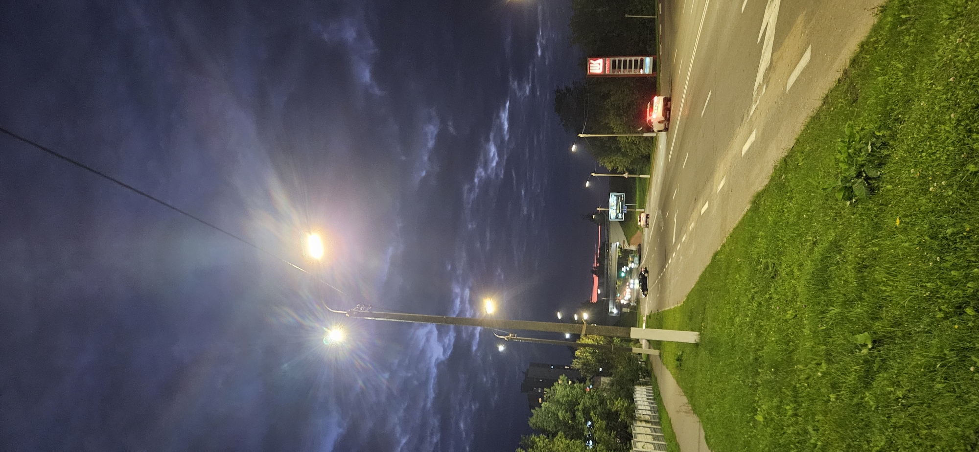 The image shows a street scene at dusk in Vitsebsk, Belarus. The sky is dark blue with heavy clouds, and streetlights are illuminated. A paved road runs through the center of the image, with a grassy median and sidewalk on the left. Several cars are visible on the road, including a white car with flashing red lights that appears to be a police vehicle. A red and white sign with Cyrillic lettering, likely a gas station or commercial advertisement, stands on the right side of the road. Buildings and trees are visible in the background. The overall impression is of a typical urban environment at the end of the day.