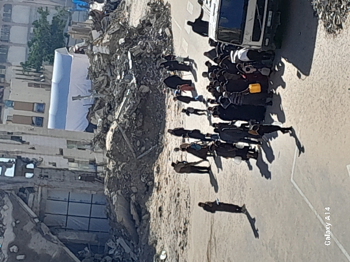 The image depicts a devastated urban street scene in Khan Yunis, Palestinian Territory. In the background, multiple multi-story buildings exhibit severe structural damage, including partial collapse, exposed interiors, and large openings, with some windows covered by white sheeting. Extensive piles of concrete, rebar, and other construction debris are scattered across the midground. A white tent with a blue roof stripe is visible amidst the rubble in the midground, to the left of center.
In the foreground, a paved road with faint white markings is present. A crowd of approximately 30-40 individuals, including men, women, and children, is clustered on the right side of the road around a white truck, partially obscured, with a visible Volvo emblem on its grille. Many individuals in the crowd are holding or carrying yellow plastic jerrycans and other assorted containers, indicating a gathering for resource distribution or collection. One person, dressed in dark clothing, stands slightly apart on the left side of the road, looking down at an object in their hands. Strong sunlight casts long, distinct shadows across the scene, particularly from the individuals and the truck. The overall environment conveys widespread destruction and a communal gathering for essential supplies.