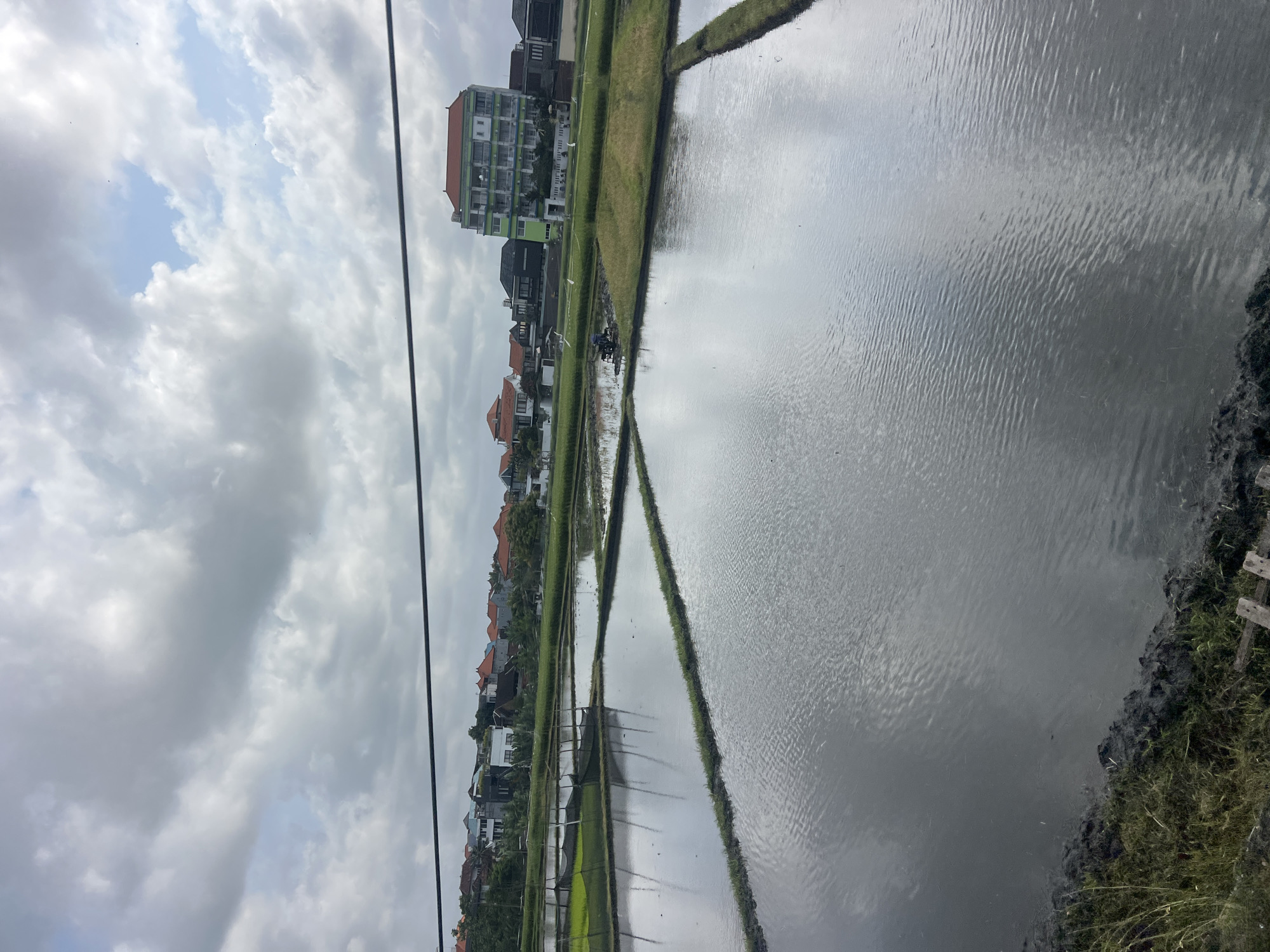 This image captures a panoramic view of flooded rice paddies in the vicinity of North Kuta, Indonesia. The foreground is dominated by the reflective surface of the water, with the dark, moist soil and lush green grass lining the edges of the paddies. In the middle ground, the landscape transitions to neatly partitioned rice fields, some appearing freshly flooded and others with a lighter green hue, possibly indicating young rice plants. A solitary figure is visible operating a small agricultural machine, likely a tiller, in one of the fields. The background reveals a cluster of buildings, including a prominent multi-story structure with a green facade and a red roof, alongside residential homes with terracotta rooftops. The sky above is a dramatic expanse of grey and white clouds, hinting at overcast conditions. A dark wire stretches horizontally across the upper portion of the frame, suggesting the presence of overhead power lines or similar infrastructure.