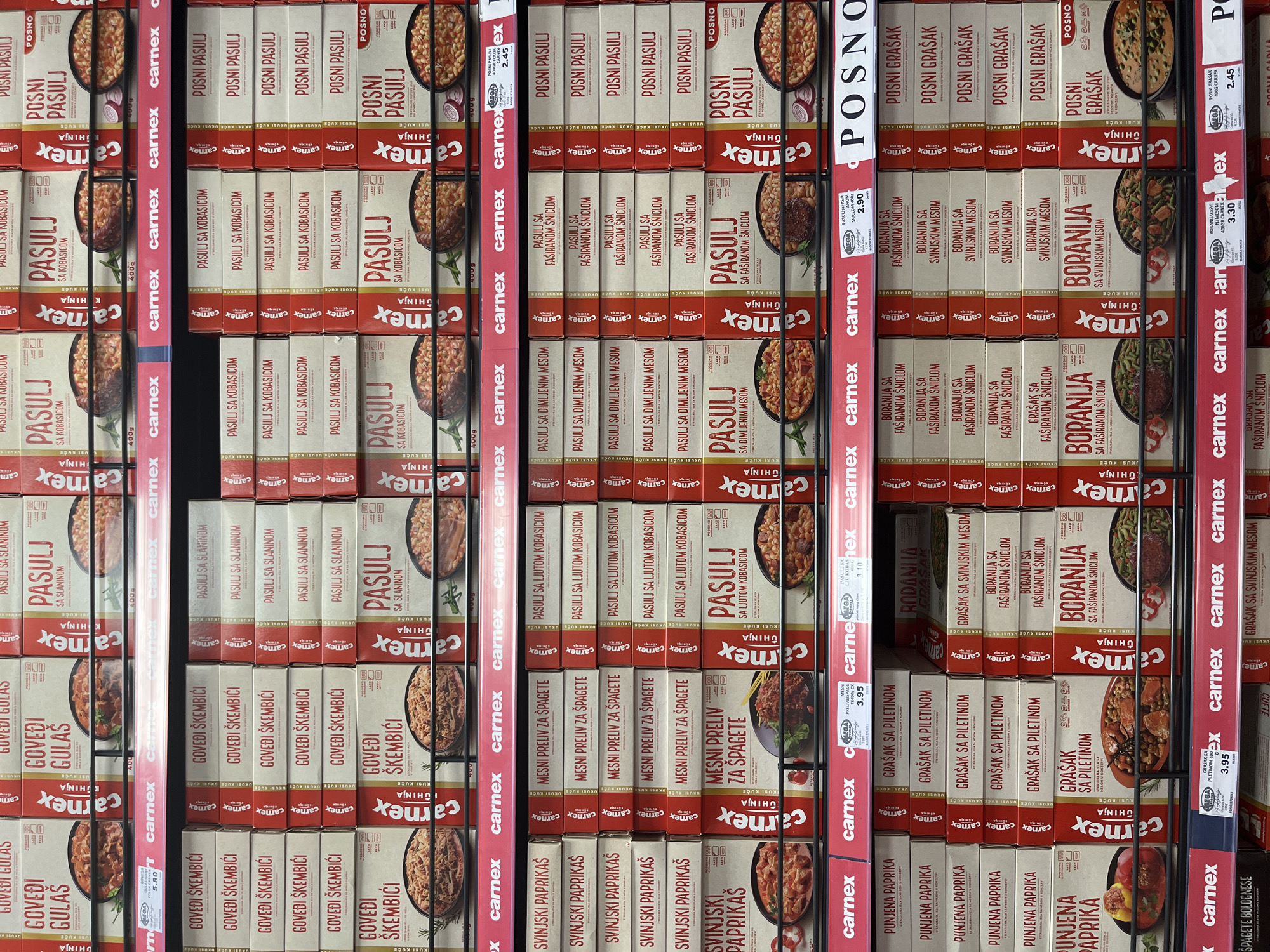 The image shows a shelf in a supermarket in Budva, Montenegro, displaying a variety of canned food products, primarily from the brand Carnex. The products include "Govedi Gulaš" (Beef Goulash), "Pasulj sa Slaniom" (Beans with Bacon), "Posni Pasulj" (Lenten Beans), "Svinjski Paprikas" (Pork Paprikas), "Mesni Preliv za Špagete" (Meat Sauce for Spaghetti), "Pasulj sa Dimnjenim Mesom" (Beans with Smoked Meat), "Pasulj sa Faširanom Snicom" (Beans with Minced Schnitzel), "Posni Grašak" (Lenten Peas), "Punjena Paprika" (Stuffed Pepper), "Grašak sa Piletinom" (Peas with Chicken), "Rodanija Grašak" (Rodanija Peas), "Boraniija sa Faširanom Sniclom" (Boraniija with Minced Schnitzel), and "Boraniija sa Svinjskim Mesom" (Boraniija with Pork Meat). The products are arranged in rows and columns on black metal shelves, with red Carnex branding visible on the packaging and on some of the shelf edges. Some products have price tags, indicating they are for sale. The lighting appears to be from overhead supermarket lights, creating a bright and even illumination. There are no people visible in the image.