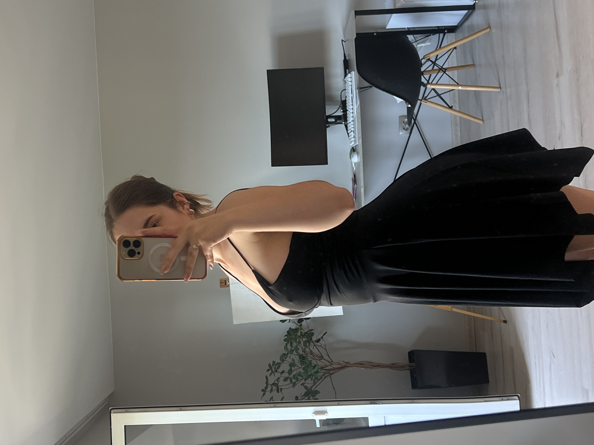 A woman is taking a mirror selfie in what appears to be a modern apartment. She is wearing a black slip dress and is holding a smartphone in her right hand. The phone has a gold case and a prominent camera array. The woman has light brown hair pulled back and is wearing gold hoop earrings and a ring on her left hand.
The background shows a grey wall, a desk with a computer monitor and keyboard, and a black chair with wooden legs. A plant in a pot is visible in the foreground, reflected in the mirror. The overall lighting suggests daytime. The image orientation is rotated 90 degrees counterclockwise. There is no visible text in the image.