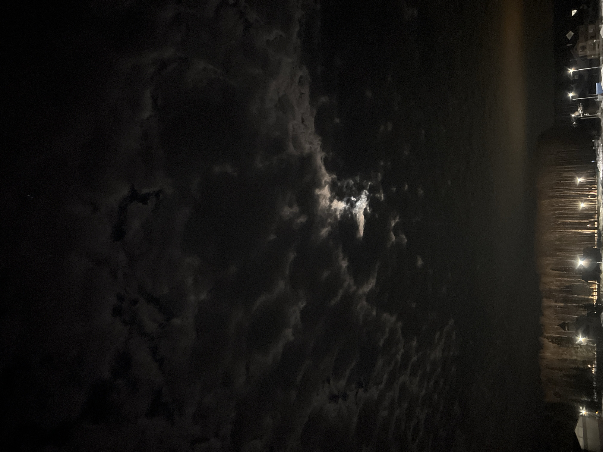 The image shows a night scene in Noviy, Russia. The sky is mostly dark with heavy, textured clouds obscuring the view. The moon is partially visible through a break in the clouds, casting a faint glow. The horizon is visible, with a cluster of lights indicating a residential or commercial area. Streetlights are visible, casting pools of light on the ground. There are no discernible people, objects, or specific actions captured.