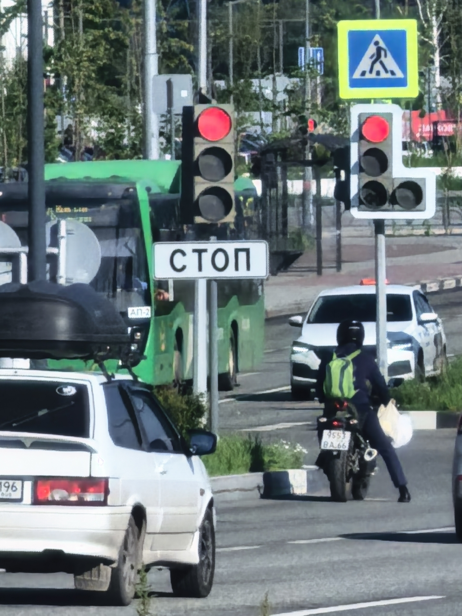 The image depicts a street scene with several vehicles and traffic signals. In the foreground, a white car is visible from the rear quarter, with its license plate partially obscured. To the right of the white car, a motorcycle with a rider wearing a green backpack is stopped at a traffic light. The rider is carrying a white bag. Further in the background, a white car is parked. A green bus is also present, with its side visible. There are traffic lights, street signs including a pedestrian crossing sign, and trees lining the street. The scene appears to be at an intersection.