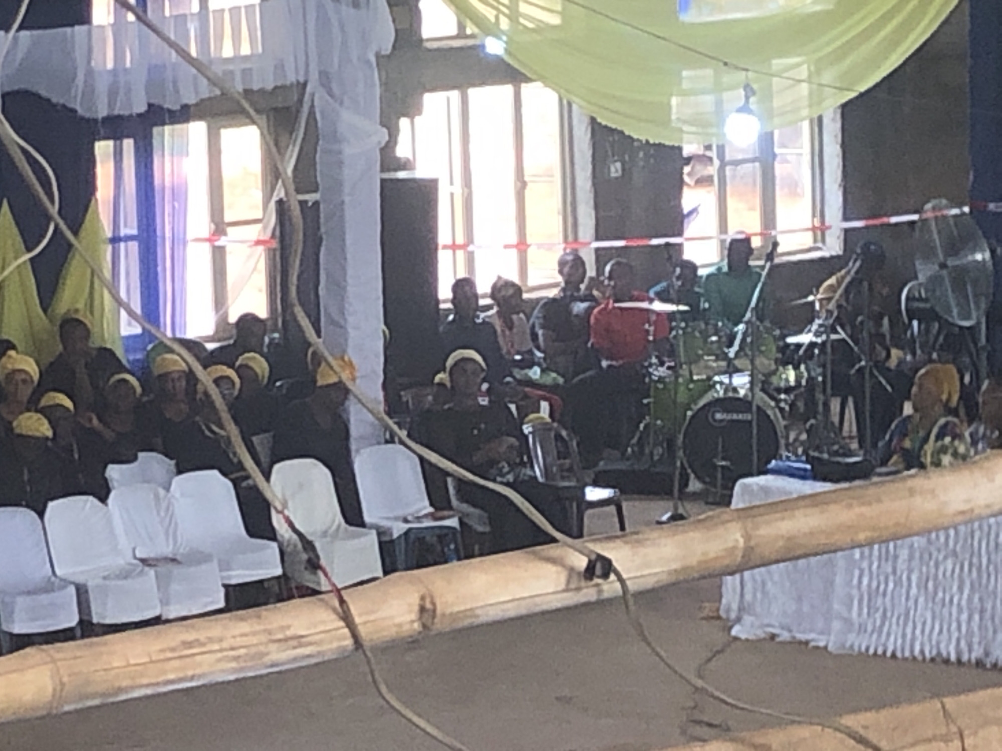 The scene depicts an event in Nteje, Nigeria. A group of people, most wearing yellow head coverings, are seated on white chairs. Some are wearing dark clothing, while others are wearing red or patterned attire. A band is set up to the right, including a drum kit, guitar, and a fan. The room has windows and is decorated with white and yellow fabric. A bamboo structure is prominently featured in the foreground.