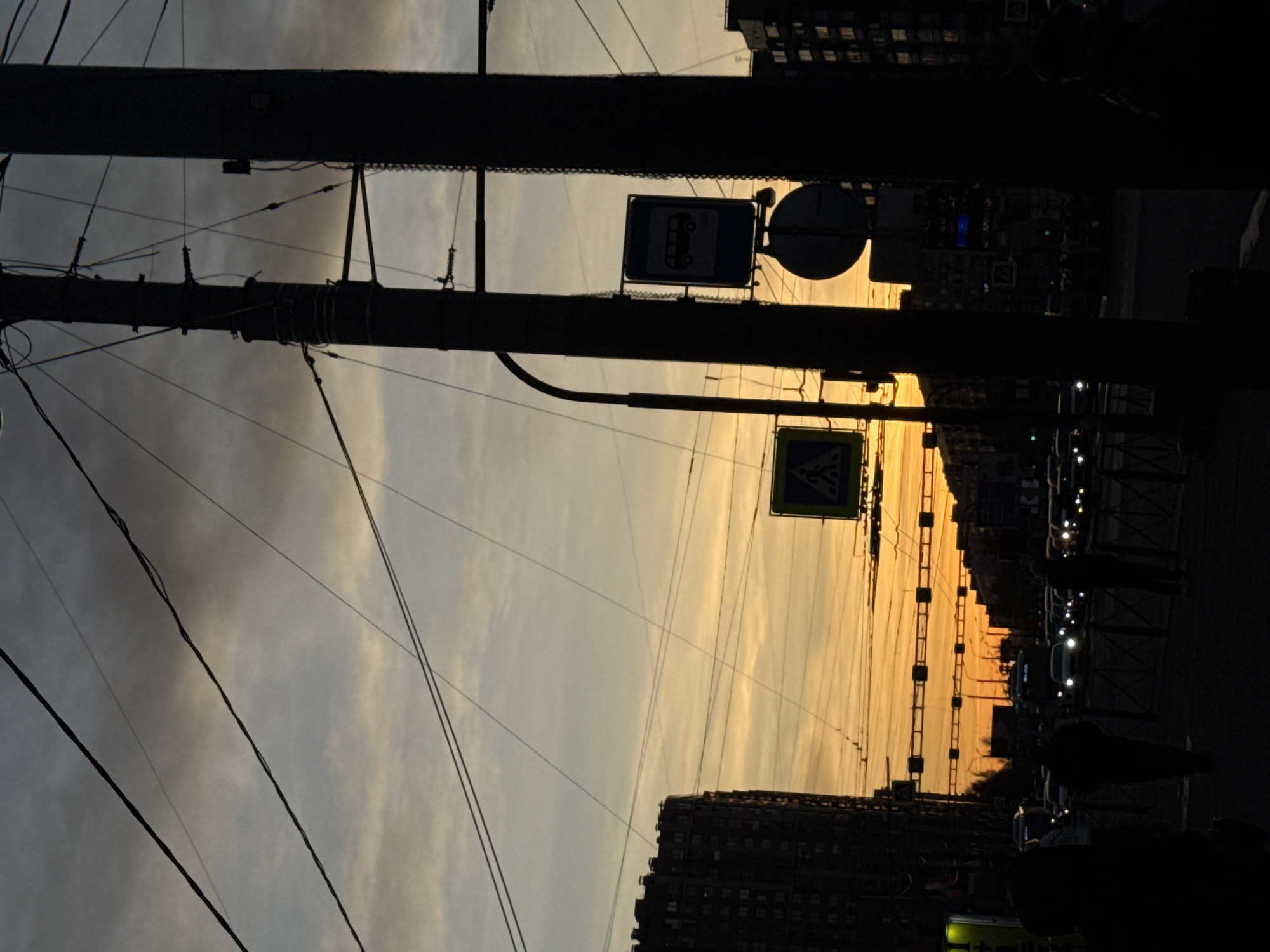 An urban street scene is captured during either sunrise or sunset, characterized by a dramatic backlighting effect that renders most elements in silhouette. The upper portion of the image is dominated by a vibrant sky, transitioning from intense golden and orange hues at the horizon to lighter grays and blues higher up, with scattered clouds.
In the foreground, a large utility pole, dark and silhouetted, rises vertically on the left, supporting a complex network of overhead power lines or tram wires that crisscross the sky. Another pole is partially visible behind it.
Mid-ground features include several prominent road signs, also seen in silhouette against the bright sky. One is a rectangular blue sign with a white bus icon, likely indicating a bus stop or lane. To its right, a circular sign is partially visible. Further right and lower, a yellow triangular sign with a black pedestrian crossing symbol is present.
Along the horizon, the dark silhouettes of multi-story buildings line the street, interspersed with trees. At street level, the faint outlines of moving vehicles are visible, with their headlights and taillights glowing against the darkness. The silhouettes of a few pedestrians can also be seen on the right, suggesting activity on the sidewalk.
The overall impression is one of a bustling city transitioning between day and night, with the urban infrastructure and human activity cast against a beautiful, naturally lit sky. There is no legible text visible in the image.