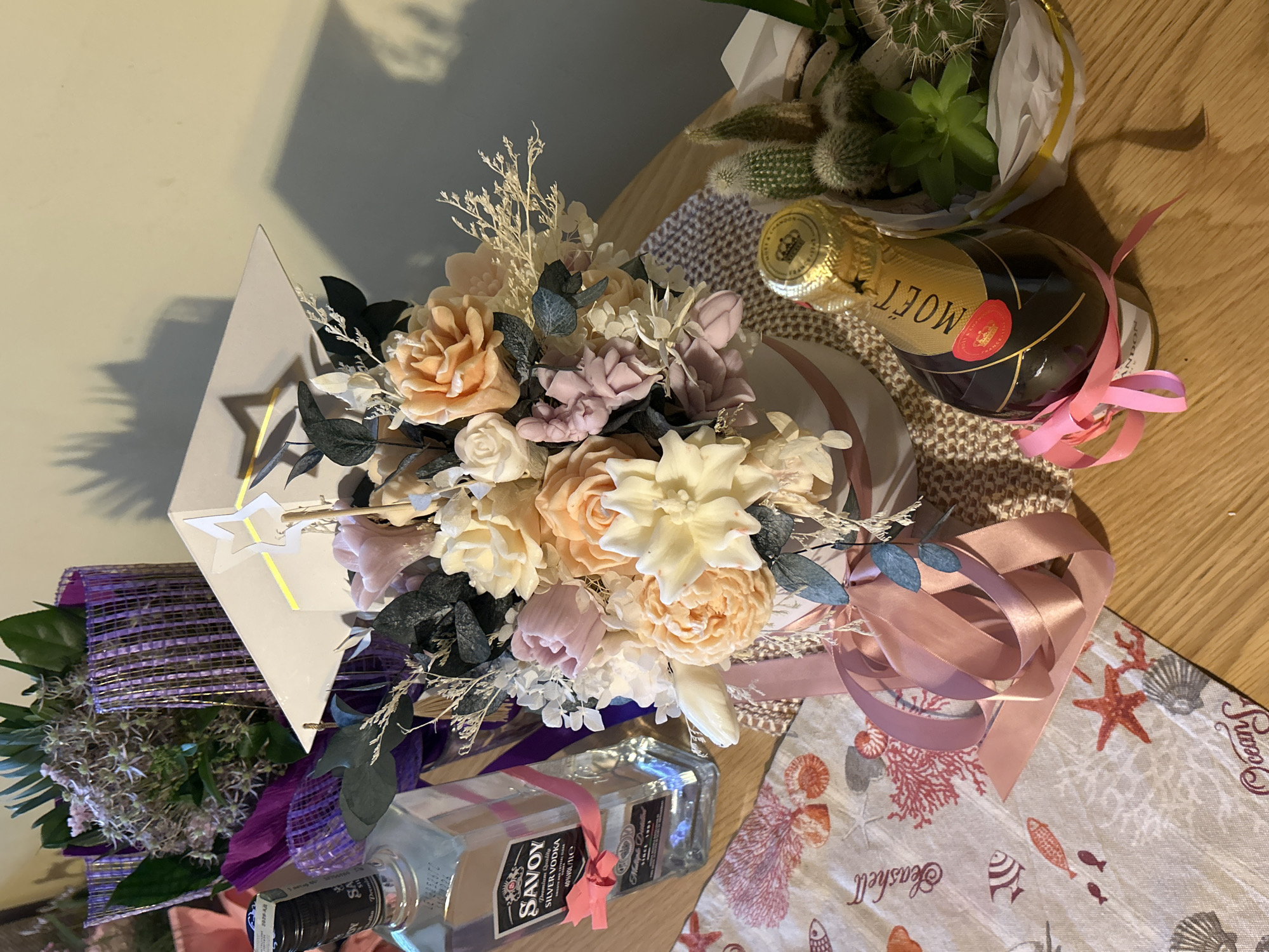 An overhead shot captures a celebratory arrangement of gifts on a light-colored wooden table. The central focus is a large bouquet of artificial or preserved flowers, featuring blooms in soft shades of peach, cream, white, and pale lavender-pink, interspersed with dried white accents and dark green/blue foliage. The bouquet is presented in a light grey box with a star-shaped ornament and gold stripe.
To the right of the flowers, a small bottle of Moët & Chandon champagne, with its characteristic gold foil and red seal, rests on a textured fabric and is adorned with a pink ribbon. Above it, a potted plant containing several cacti and succulents is wrapped in white and gold paper.
In the lower left, a rectangular glass bottle of "SAVOY SILVER VODKA," labeled "Premium Quality" and "Multiple Distilled," is tied with a matching pink ribbon. A partially visible bouquet wrapped in purple mesh with dried purple flowers is also present in this area. A patterned fabric beneath some items features an "Ocean" and "Seashell" motif with illustrations of marine life like shells, starfish, and fish. The overall scene suggests a gift-giving occasion or celebration. Shadows are cast on a plain wall in the background, indicating indoor lighting.