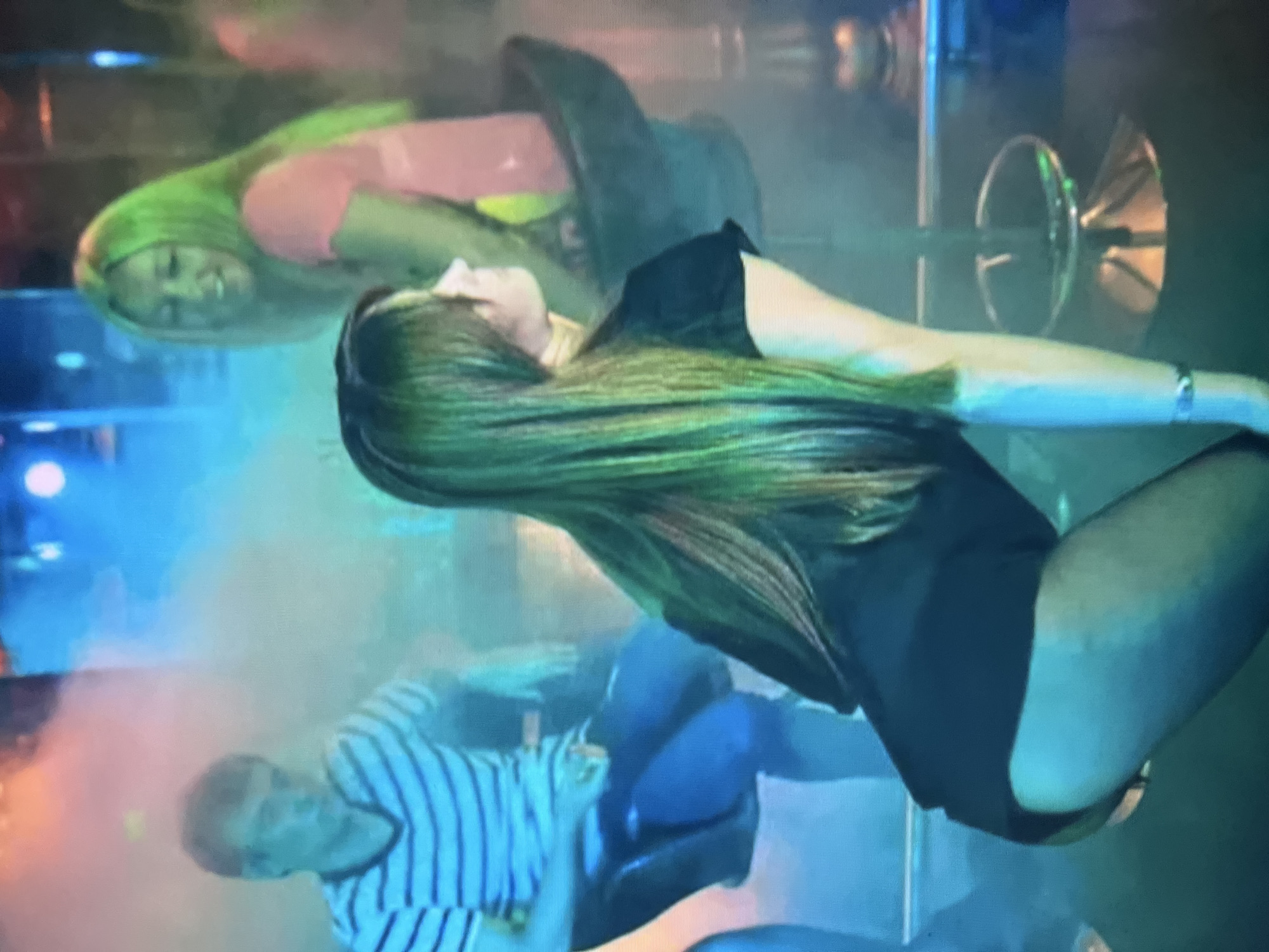 An indoor scene features three individuals in a dimly lit environment with colorful lighting and smoke. In the foreground, a person with long, dark hair is bent forward, their back to the camera, wearing a black top and what appear to be black tights. To the left, a male with short, light brown hair sits on a stool, wearing a striped t-shirt and dark pants. In the background, a person with long, bright green hair smiles and wears a pink top, seated on a stool. Bar stools are visible, as is a pole. The scene appears to be a nightclub or similar entertainment venue. The location in the city of Qarağandı city administration, Kazakhstan is not discernable from the image.
