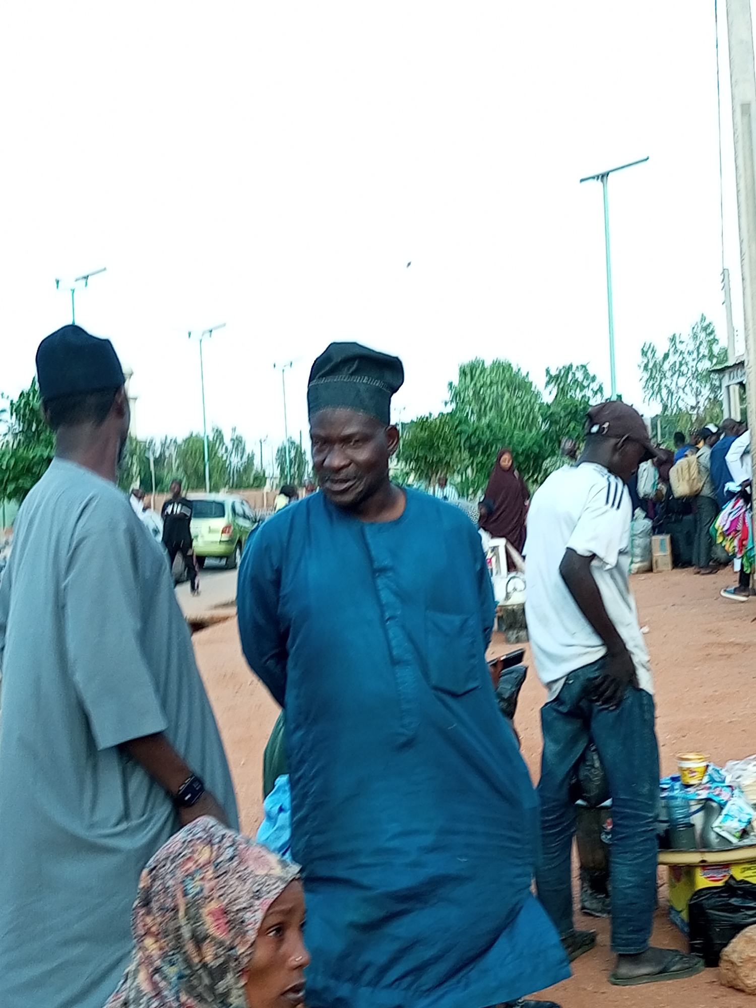A group of people are gathered outdoors, appearing to be in a market or a public gathering area. The scene is dominated by individuals in traditional Nigerian attire, suggesting a cultural context. The environment appears to be dusty with some vegetation in the background, and light poles are visible, indicating an urban setting. The people are dressed in long robes and head coverings, typical for this region. Some individuals are looking at each other, engaged in conversation, while others are focused on different activities. The overall atmosphere seems relaxed and casual, with a sense of community present. The presence of stalls with goods suggests commercial activity. The image appears to be taken in Gombe, Nigeria, a city known for its vibrant markets and cultural diversity.