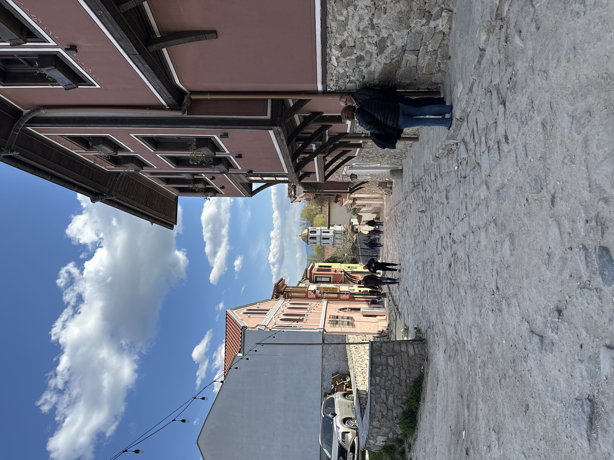 A street view in the city of Plovdiv, Bulgaria. The narrow, cobblestone street is lined with traditional-style buildings, painted in shades of pink and beige. The architecture features dark wood accents. A couple leans against the building wall, and other people walk further down the street. A white bell tower is visible at the end of the street. On the left side of the street, there is a white building, and a car is parked nearby. The sky is blue with fluffy white clouds.