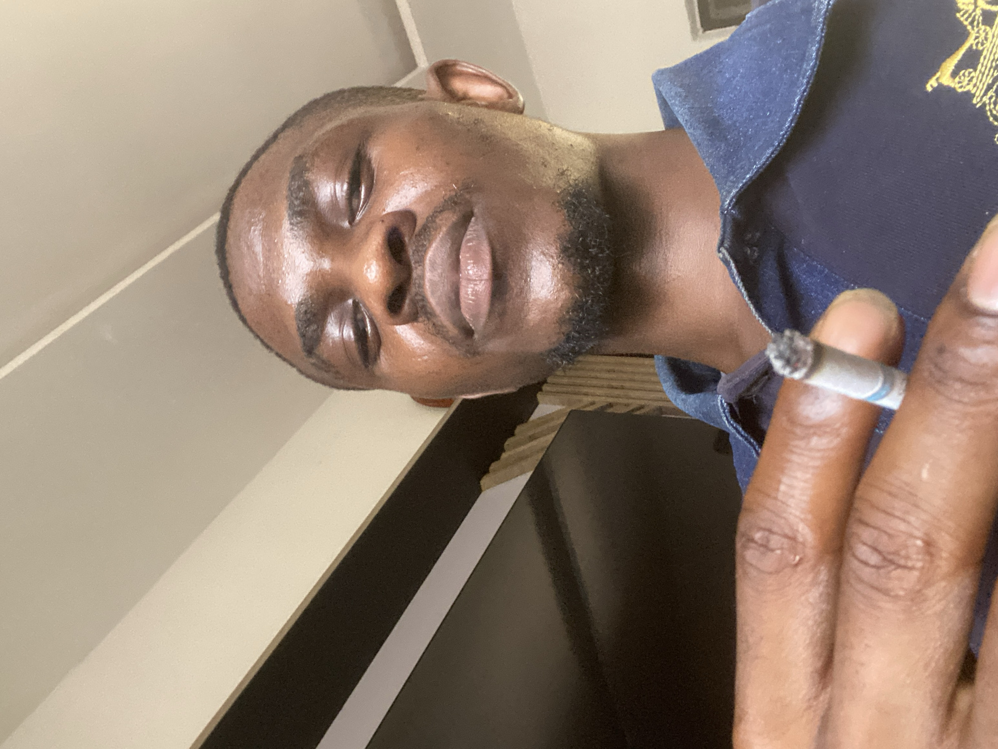 A dark-skinned man with a short haircut and beard is holding a lit cigarette. He's wearing a blue shirt. The background includes a beige wall and a black cabinet. It is not possible to confirm the location in Abuja, Nigeria from this image alone.