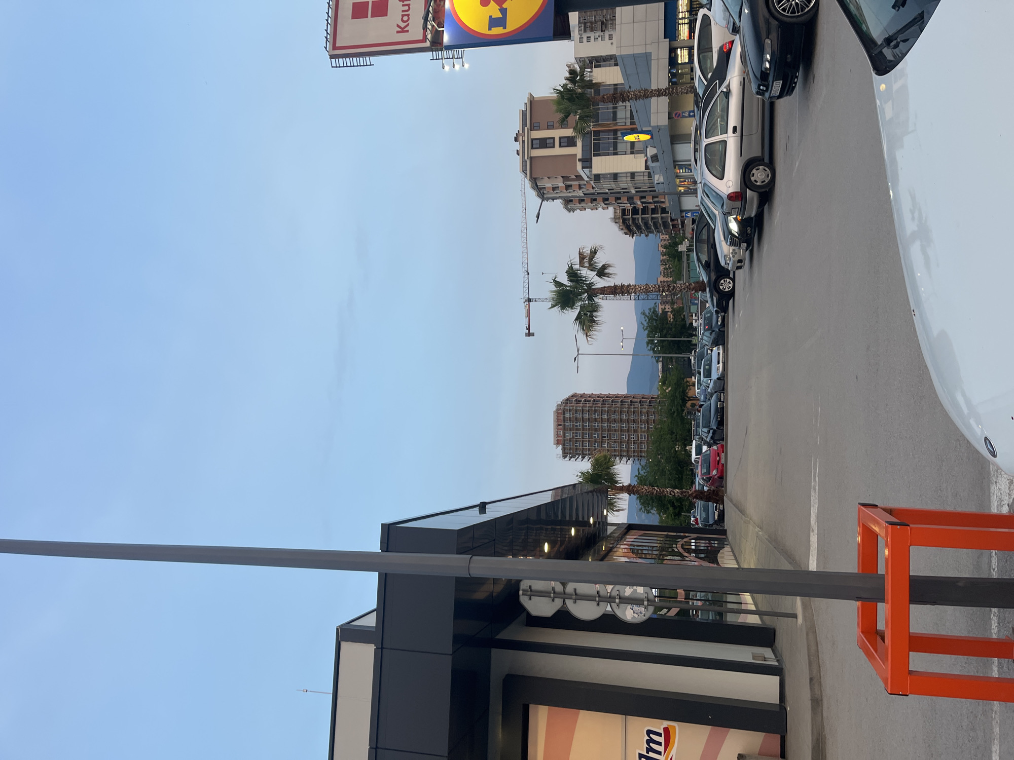 The image depicts an exterior scene at a shopping center in Plovdiv, Bulgaria, during the late afternoon or early evening. The sky is a pale blue with some faint cloud cover. In the foreground, a portion of a white car is visible, along with a grey lamppost surrounded by an orange protective barrier. To the left, the modern facade of a building, likely a retail store, is present, with large windows and dark grey paneling.
The middle ground of the image shows a parking lot filled with various cars, including a red car, silver cars, and black cars. Palm trees are interspersed throughout the scene, adding a touch of greenery. In the background, several multi-story buildings are visible, some appearing to be under construction, indicated by a construction crane. A large sign for "Lidl" is prominently displayed on the right side of the image, along with another sign that reads "Kaufland" above it. The overall environment suggests a commercial area with a mix of modern architecture and some ongoing development. There are no people clearly visible in the image.