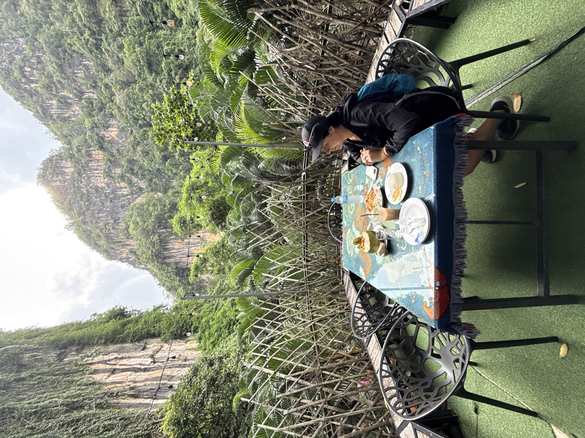 A person sits at a table in an outdoor dining area with a mountainous, jungle-covered background. The person, wearing a black jacket, black shorts, a black baseball cap, and a blue backpack, is seated at a table covered with a blue tablecloth featuring a world map design. A meal is laid out on the table, including rice, a coconut, and drinks. The dining area is decorated with bamboo fencing and string lights, and the ground is covered with artificial grass. The scene appears to be located in Chong Pli, Thailand, given the distinctive karst topography and lush vegetation characteristic of the region.