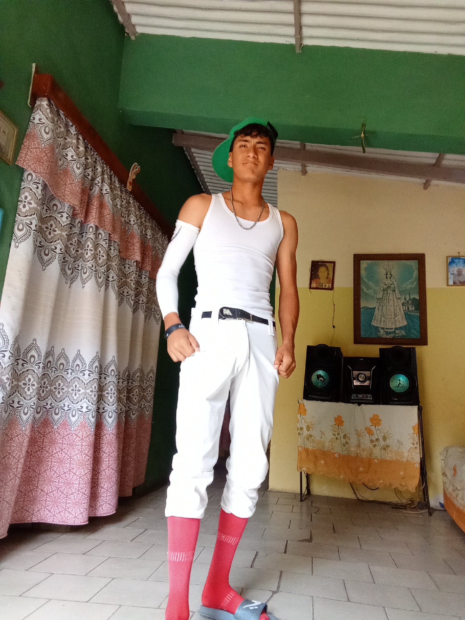 A young man stands indoors, posing for a photograph. He is wearing a green baseball cap, a white tank top, white pants, and red socks with blue sandals. A partial white sleeve covers his left arm. He also wears a silver chain necklace and a black wristwatch. The man is standing on a tiled floor, in front of a patterned curtain on the left, and a table on the right, which holds speakers and a stereo system. Above the stereo is a framed picture of the Virgin Mary. Other framed pictures are also seen on the wall. The walls of the room are painted in shades of green and yellow. The location is likely a residence in Juan Griego, Venezuela.