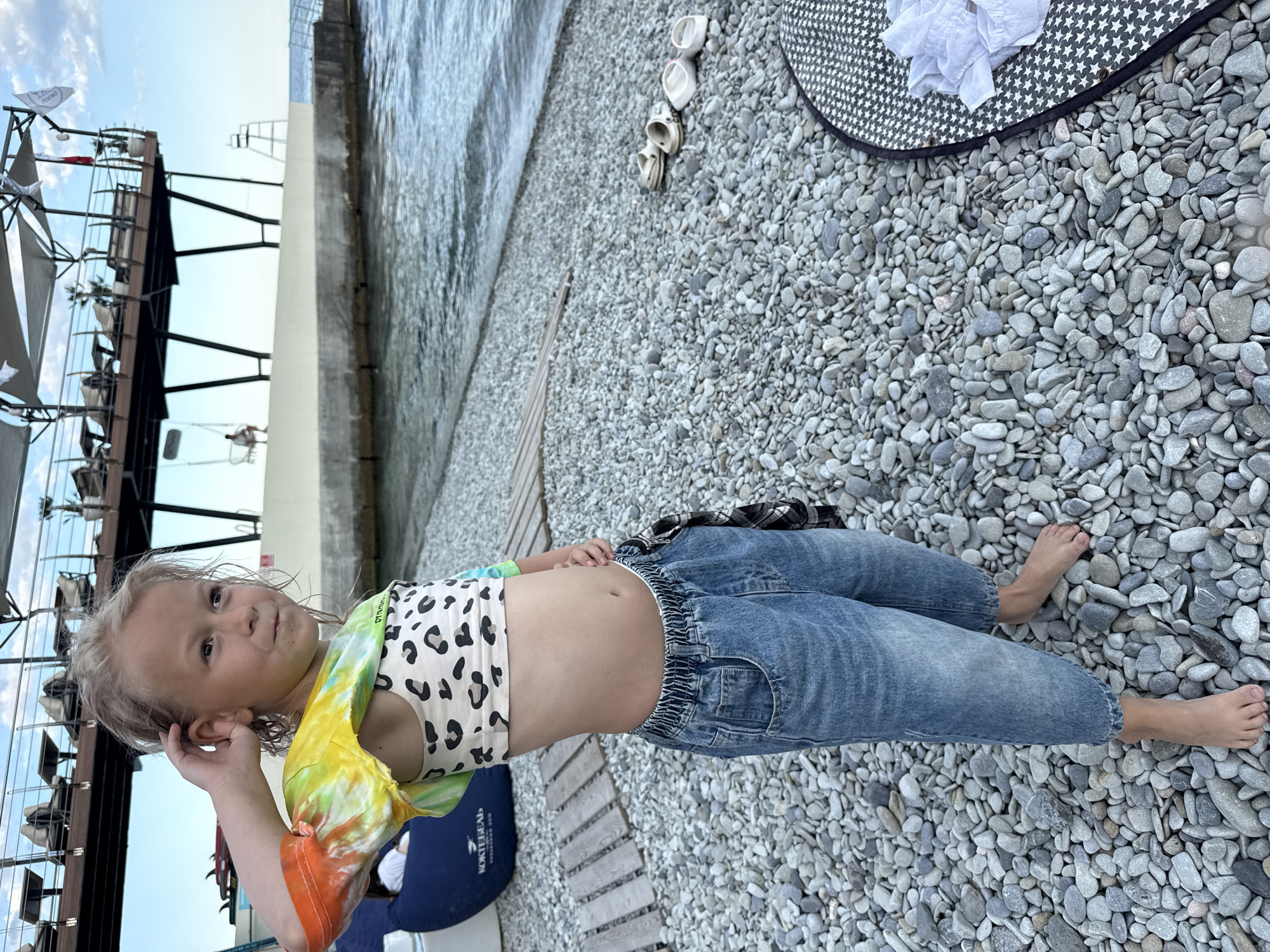 A young girl with blonde hair stands on a pebbled beach, her body angled towards the camera. She is wearing a leopard-print crop top with multi-colored tie-dye sleeves and light blue jeans. Her left hand is raised to her ear, and she has a slight smile on her face.
Behind her, the calm, gray water of the sea meets a concrete embankment. To her left, a modern structure with several levels and umbrellas suggests a beachside resort or cafe. The sky is clear with a few scattered clouds, indicating it is daytime. A large, black and white patterned hat with a white cloth draped over it lies on the pebbles to the right of the girl. Several pairs of white sandals are scattered on the ground nearby. The overall scene suggests a relaxed day at the beach.
There is a logo visible on a blue beanbag chair in the lower left of the image that appears to read "КОКТЕБЕЛЬ". The location context is provided as "город Ялта, Ukraine".